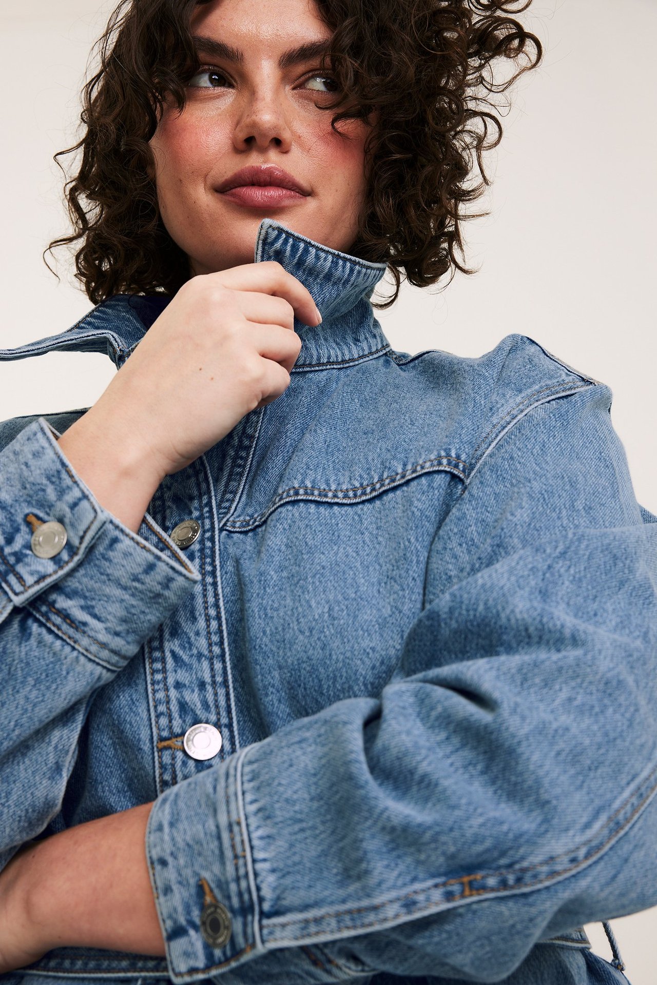 Lähikuva naisesta vaaleansinisessä denimtakissa hopeanapeilla, pitäen kauluksestaan kiinni.