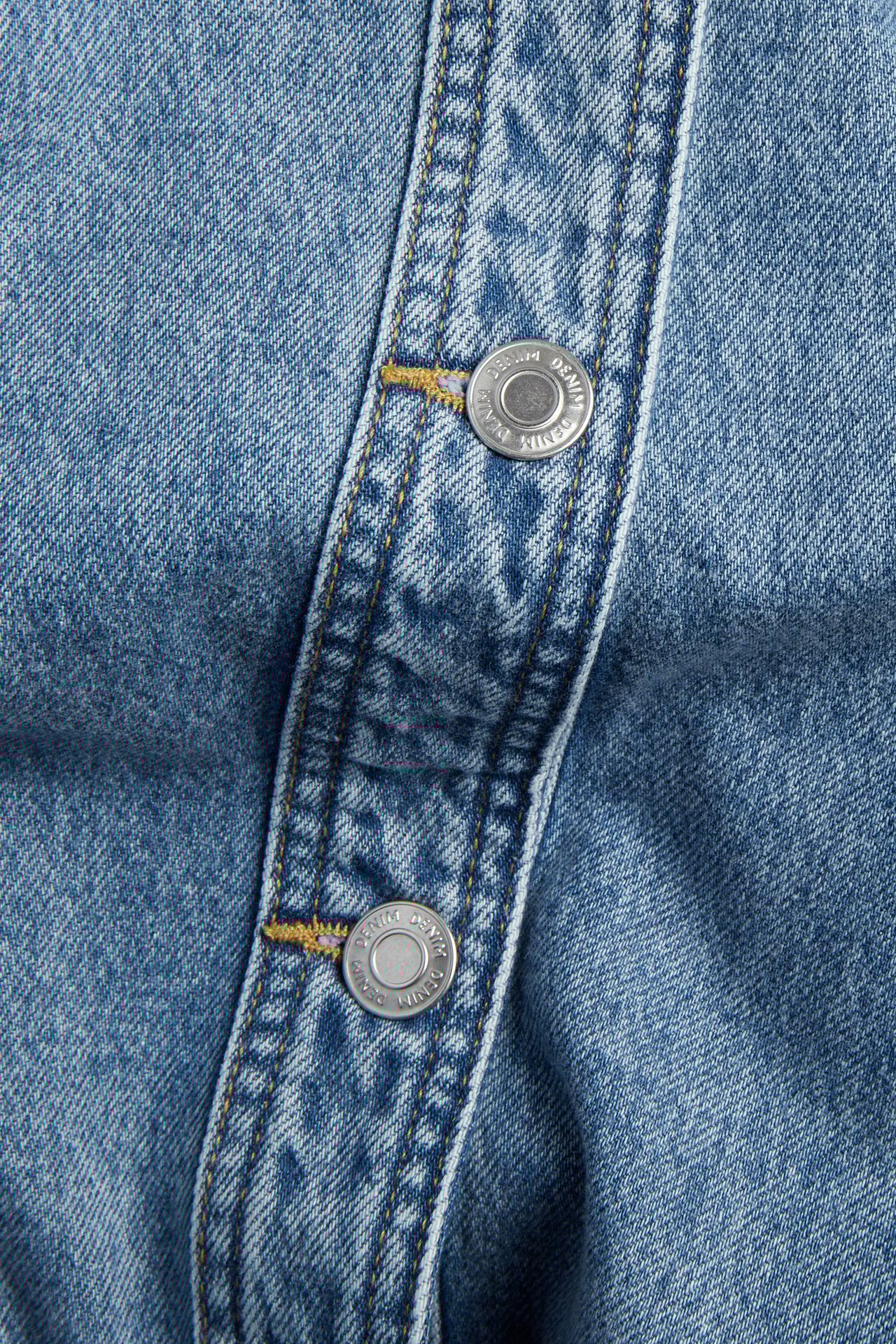 Nærbilde av lysblå dongerijakkes knappelås med to sølvknapper og gul søm.