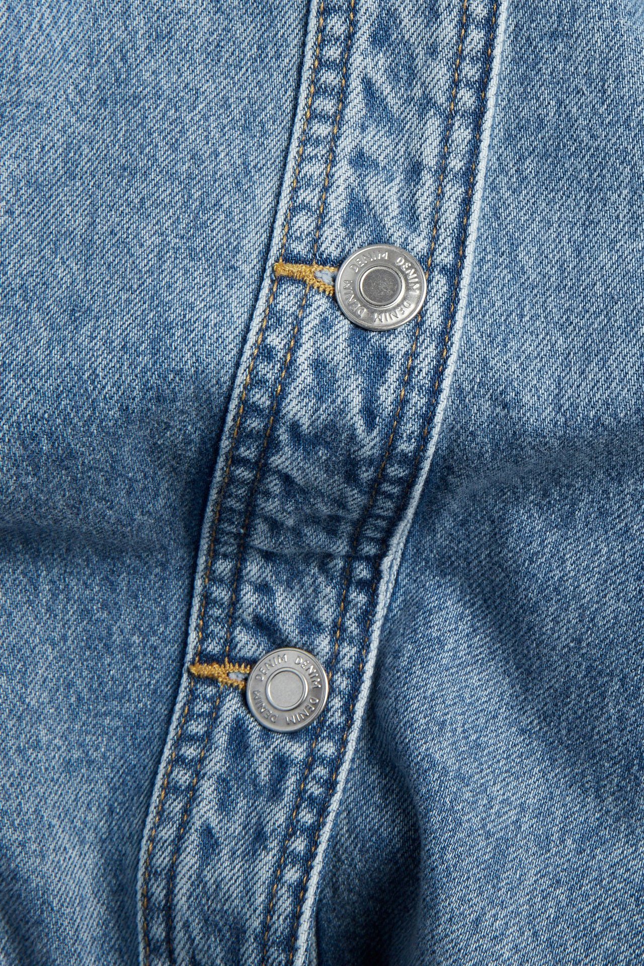 Lähikuva vaaleansinisen farkkutakin napituslistasta, jossa kaksi hopeanappia ja keltainen tikkaus.