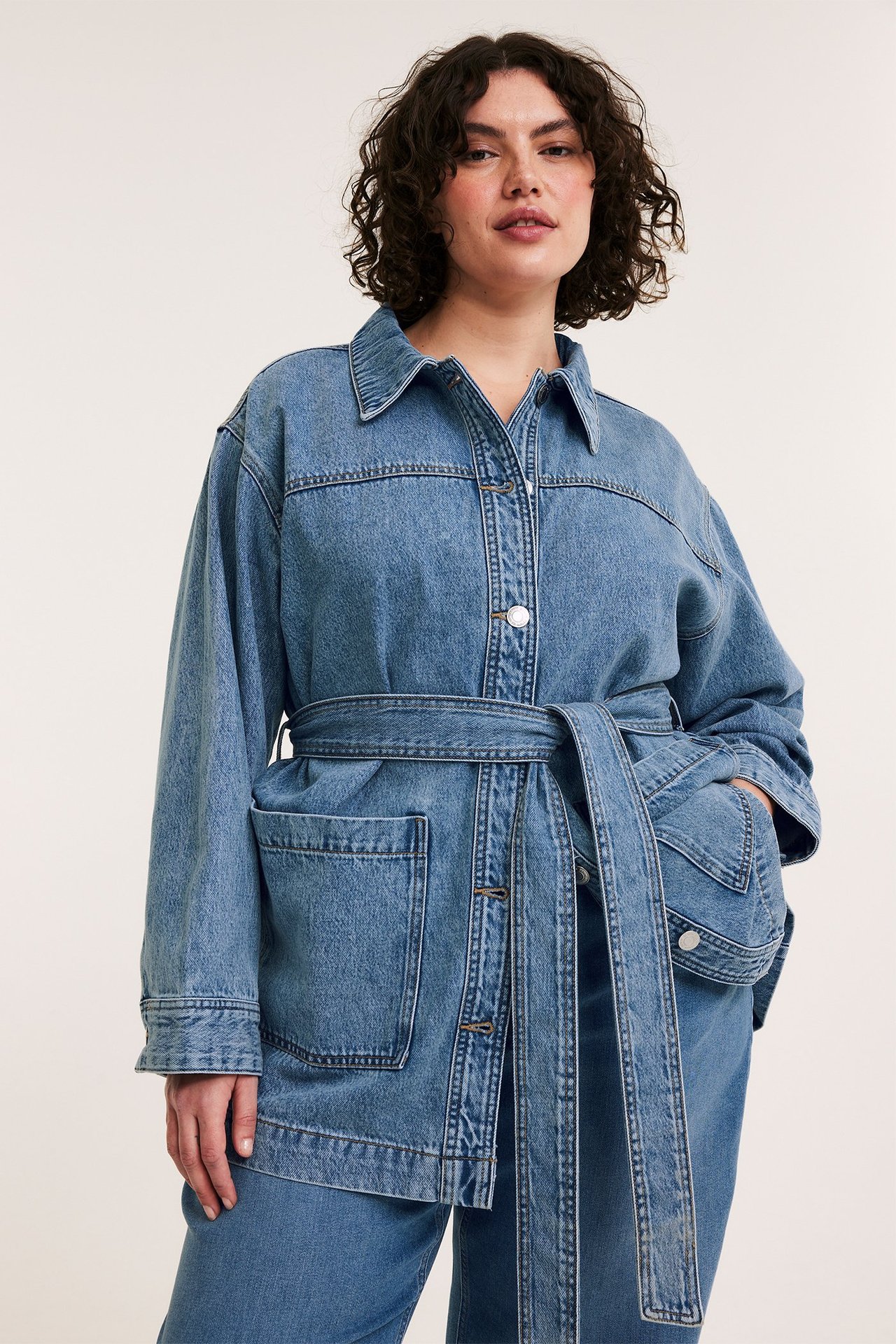 Frontutsikt av en kvinne i lys blå denimshacket med belte, lappelommer og matchende bukser.