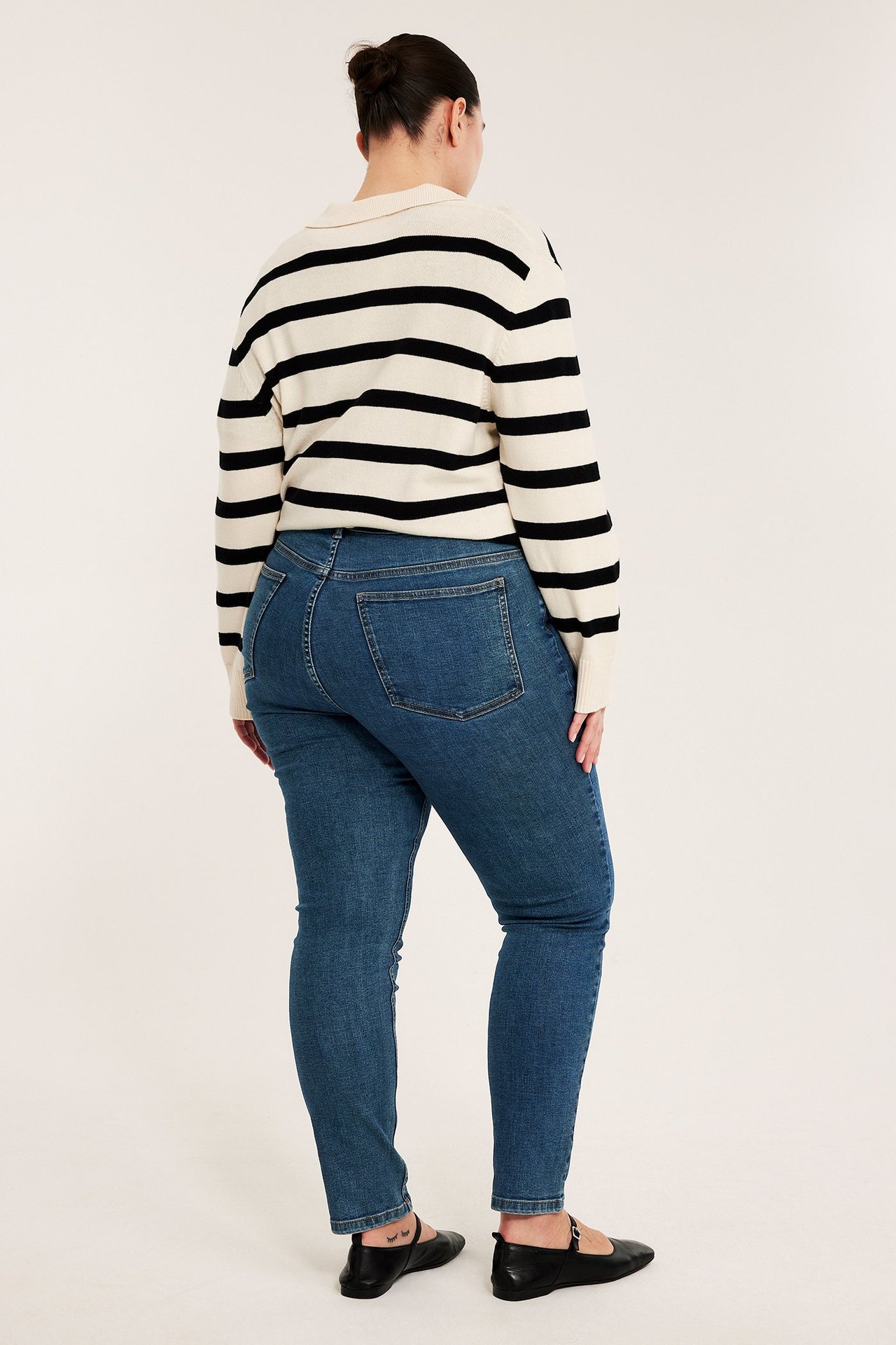 Bakifrån av en kvinna i blå slim high-waist denimjeans, en randig tröja och svarta platta skor.
