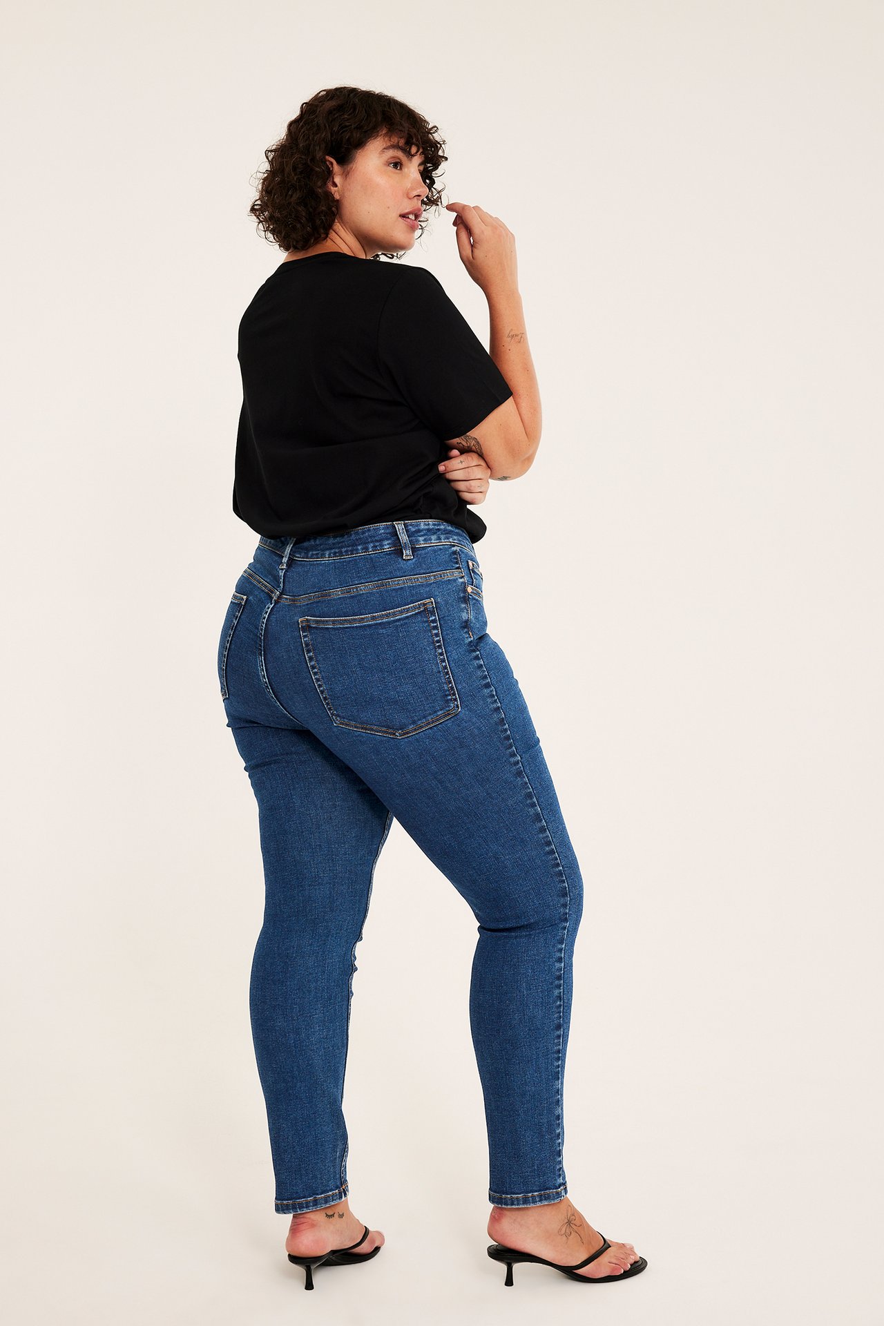 Widok z tyłu kobiety w niebieskich jeansach skinny, czarnym t-shircie i czarnych sandałach na obcasie.