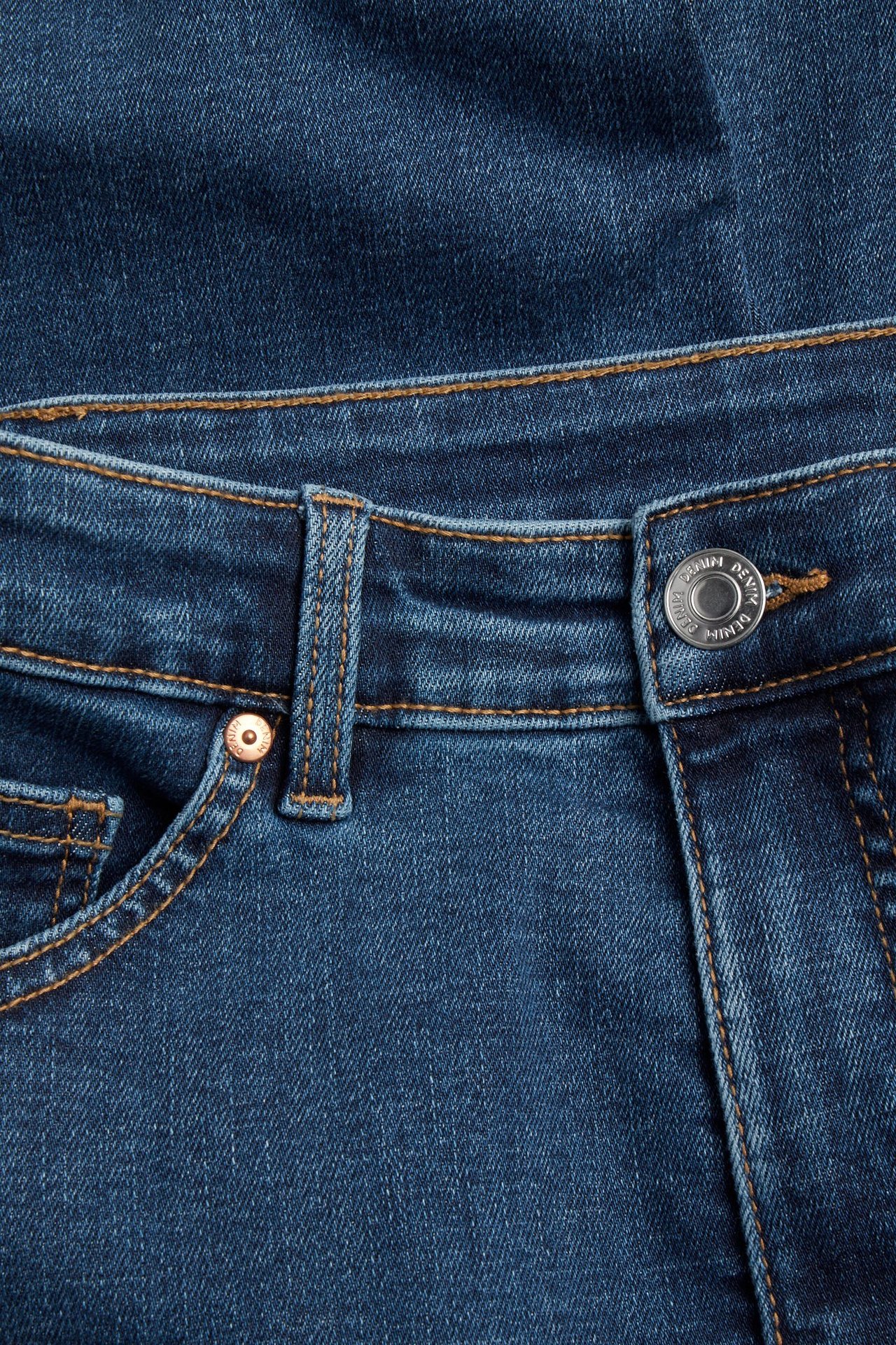 Närbild av mörkblå denimjeans som visar en silverknapp, en kopparnit och orange kontrastsömmar.