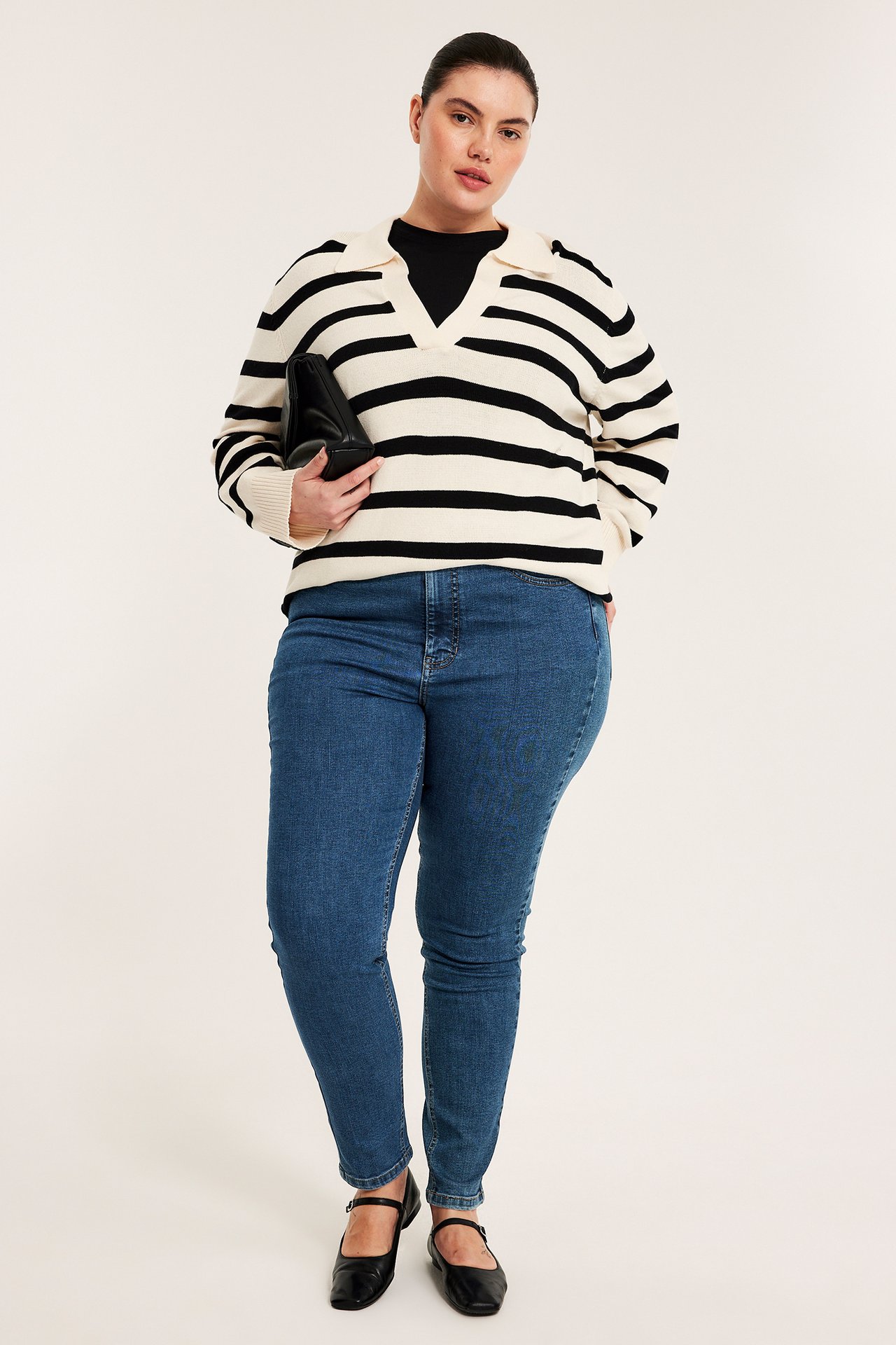 Framifrån: kvinna i blå slim high-waisted denimjeans, randig tröja, svart topp, svarta ballerinaskor, håller clutch.
