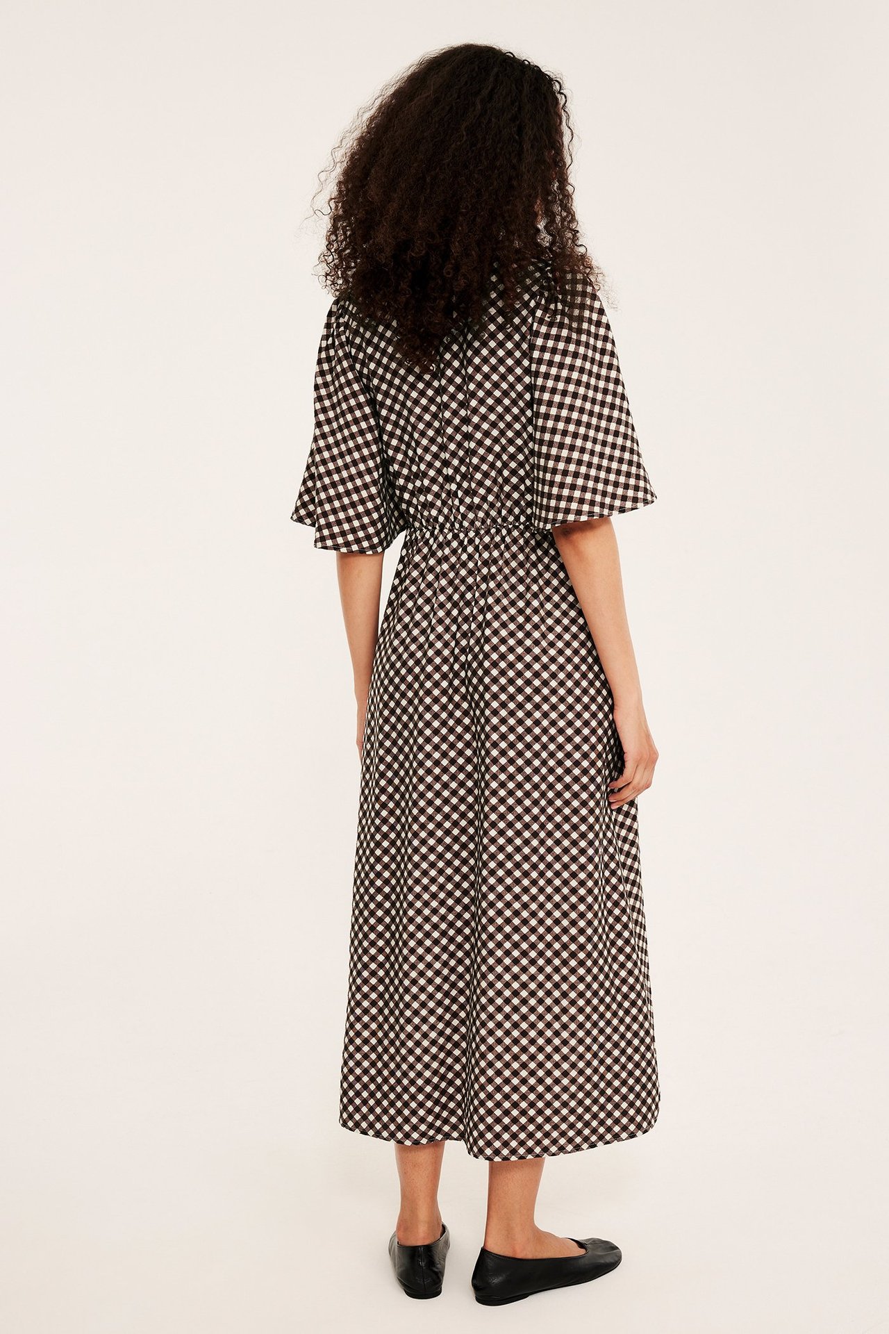 Nainen takaa, ruskea-valkoruudullinen midimekko röyhelöhihoilla ja mustat matalat kengät.