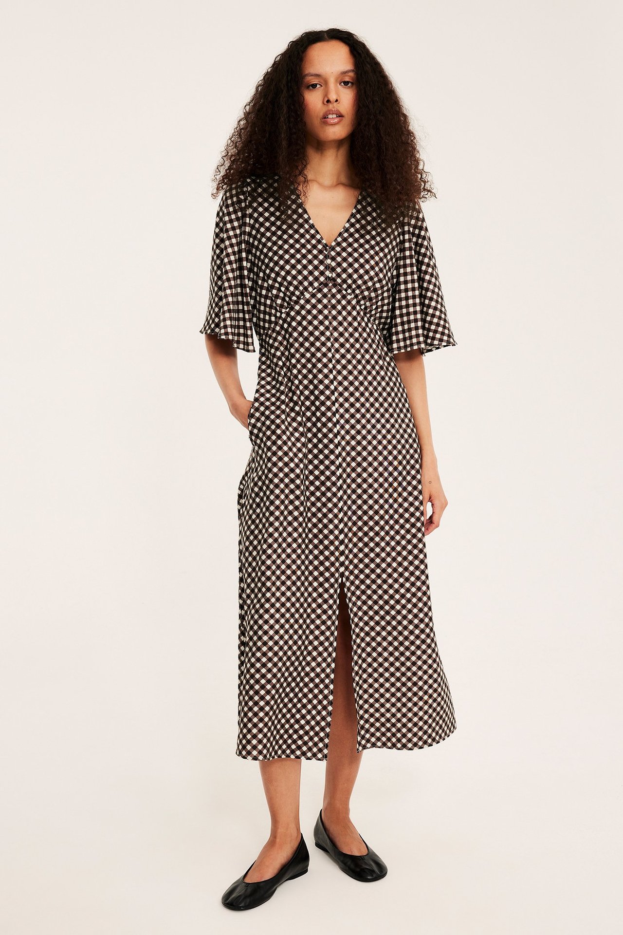 Kobieta w brązowo-białej wzorzystej sukience midi z guzikami, falbankowymi rękawami i czarnymi baletkami.