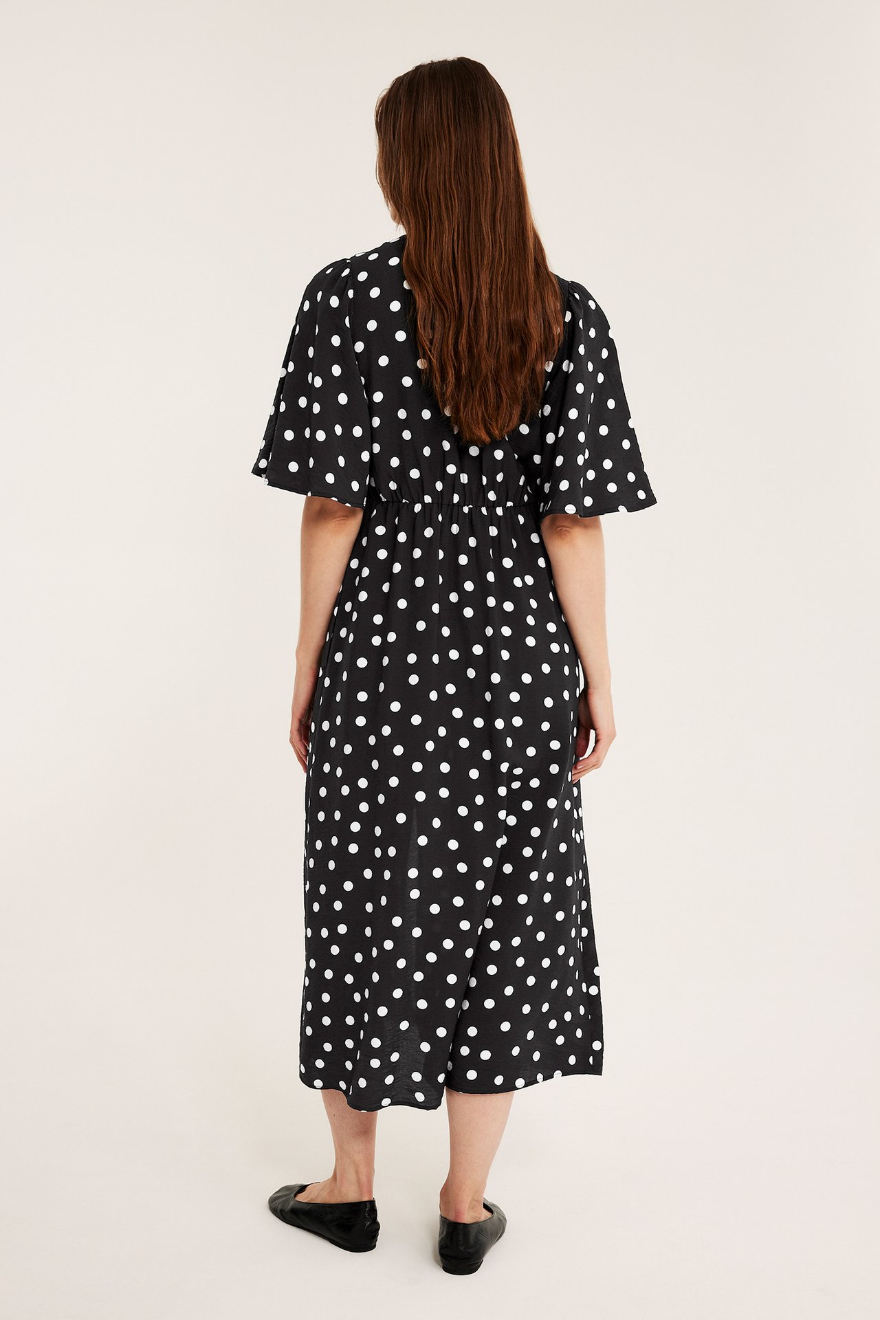 Widok z tyłu kobiety w czarnej wzorzystej sukience midi w białe kropki, z bufiastymi rękawami.