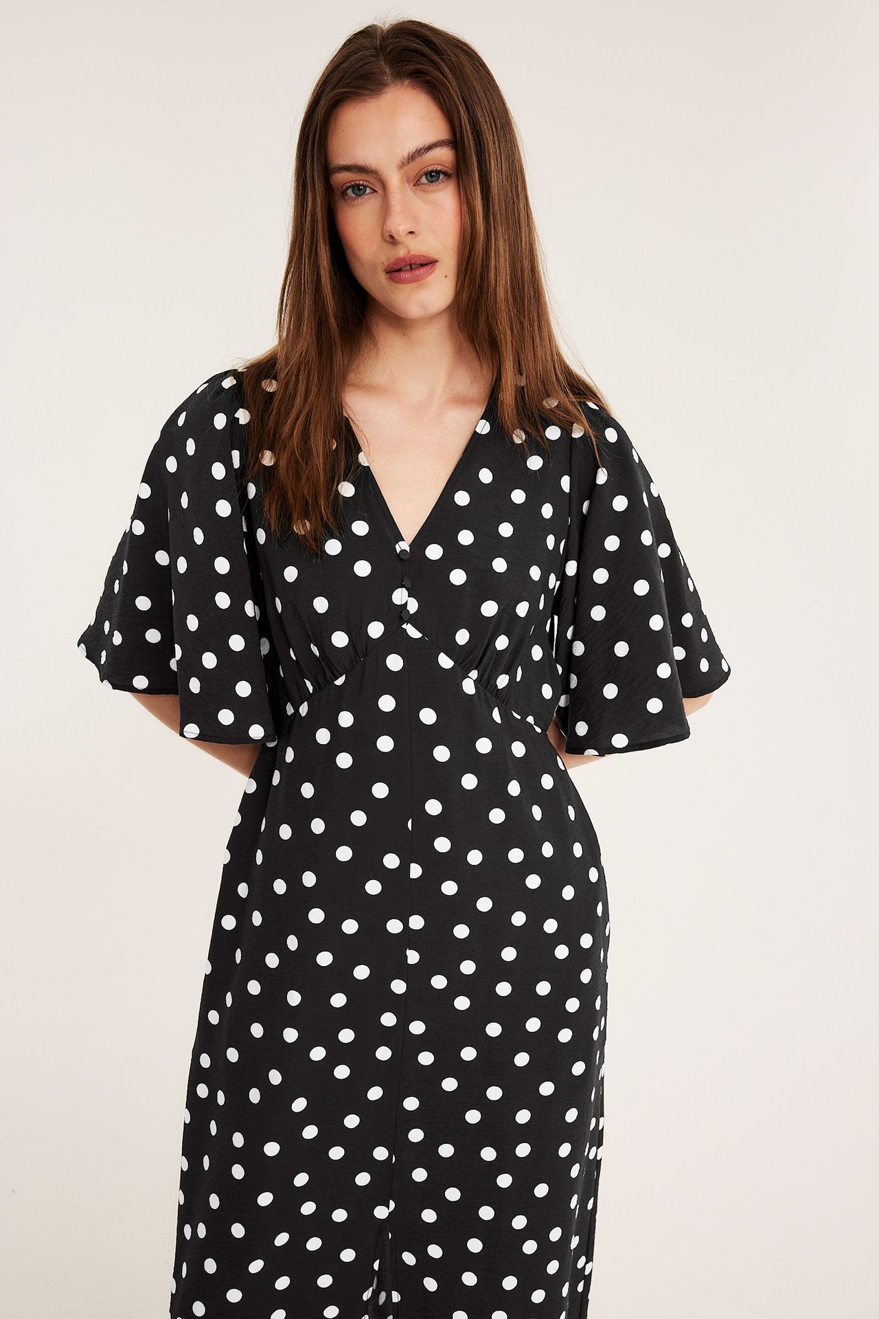 Edestäpäin: Nainen mustassa pilkullisessa midimekossa, jossa V-pääntie, napit ja röyhelöhihat.