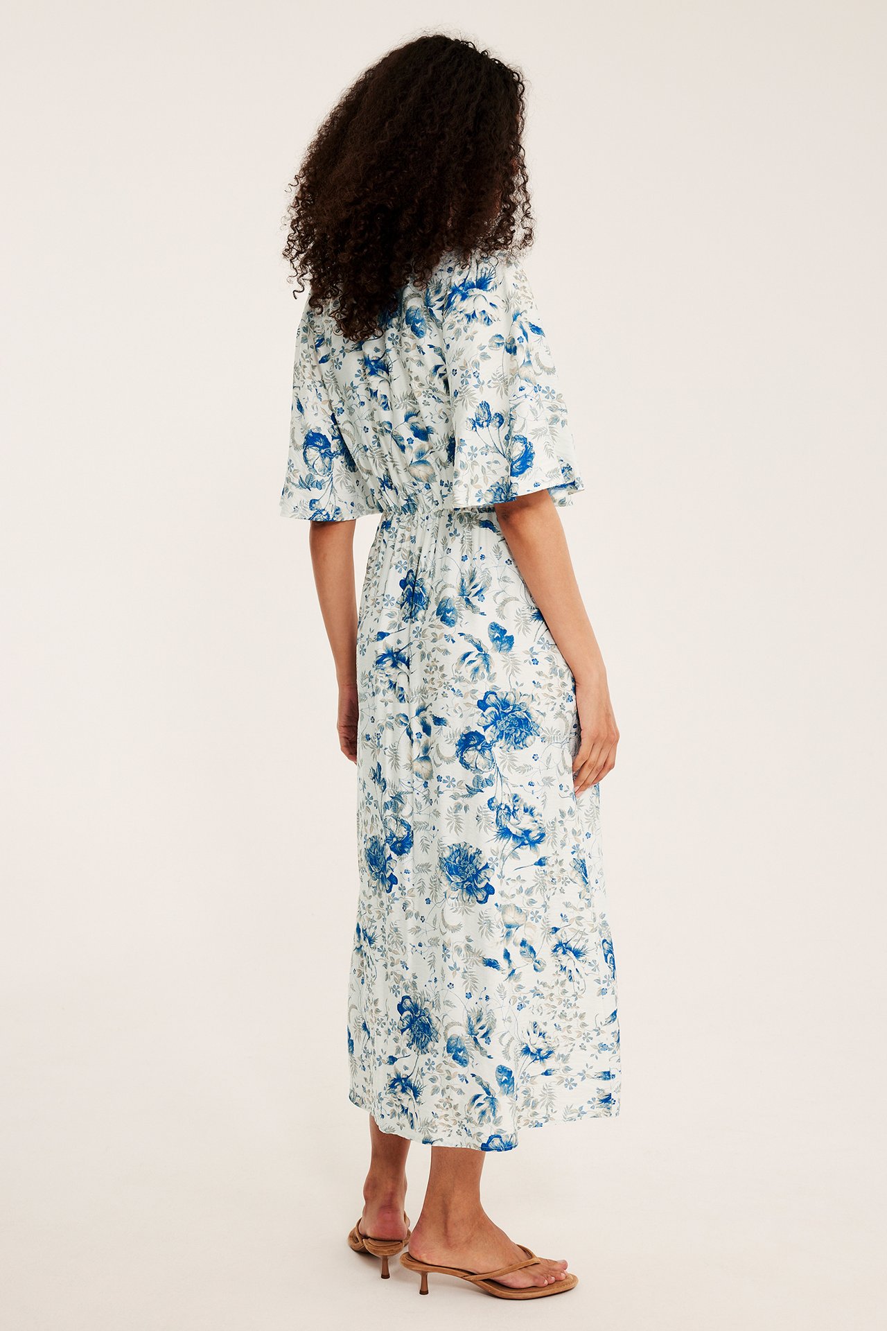 Kobieta, tyłem, biała sukienka midi, niebieskie kwiaty, falbankowe rękawy, brązowe sandały.