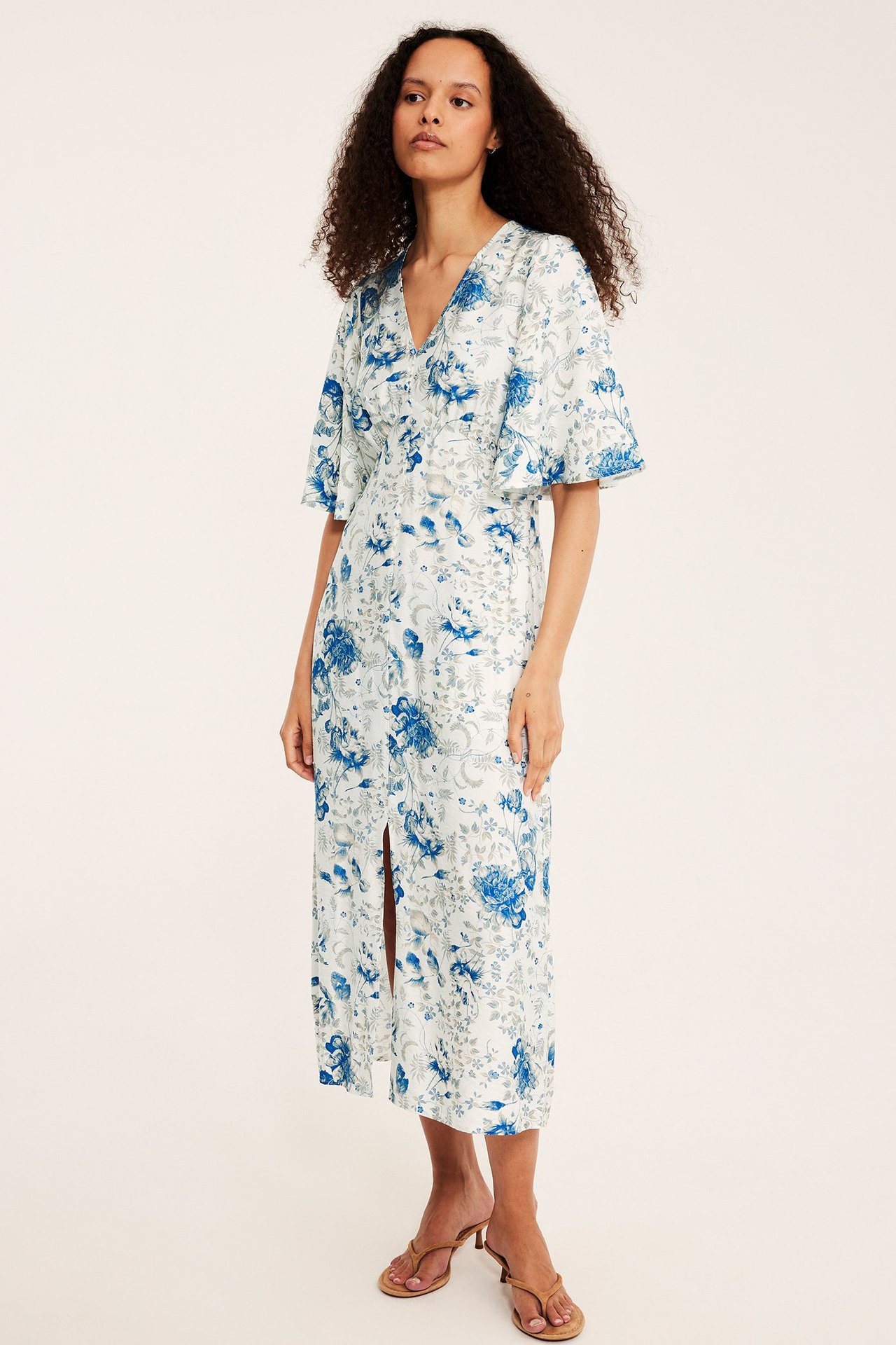 Edestä: Nainen valkoisessa sinikukkaisessa midimekossa, röyhelöhihat, napit edessä. Ruskeat sandaalit.