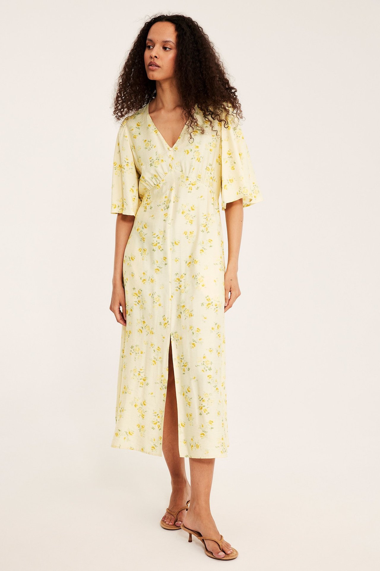 Forfra: kvinne i lys gul mønstret midikjole med V-hals, knapper og volangermer, iført brune høye sandaler.