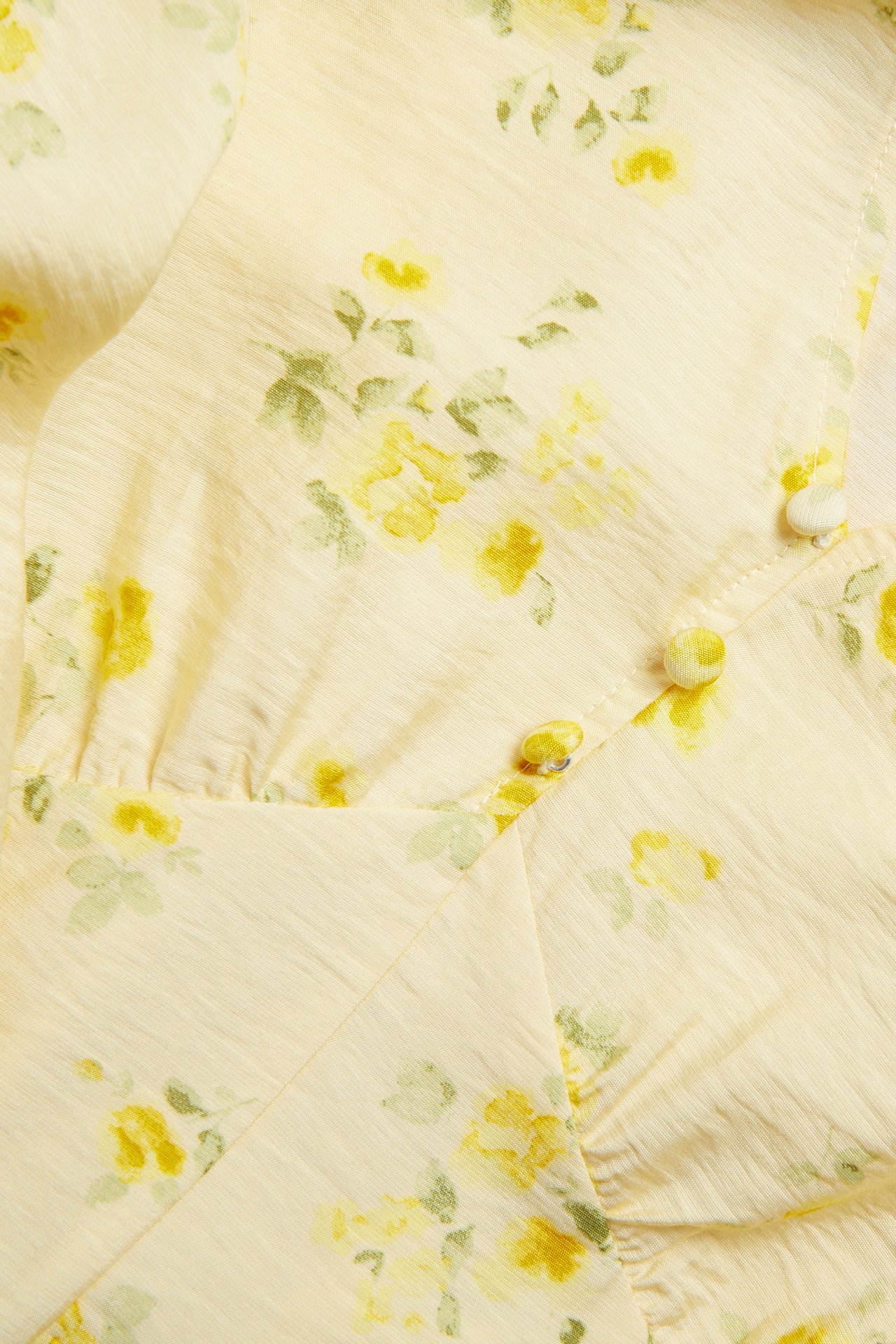 Lähikuva vaaleankeltaisesta kuviollisesta mekosta, jossa on keltainen kukkakuvio ja kangaspäällysteiset napit.