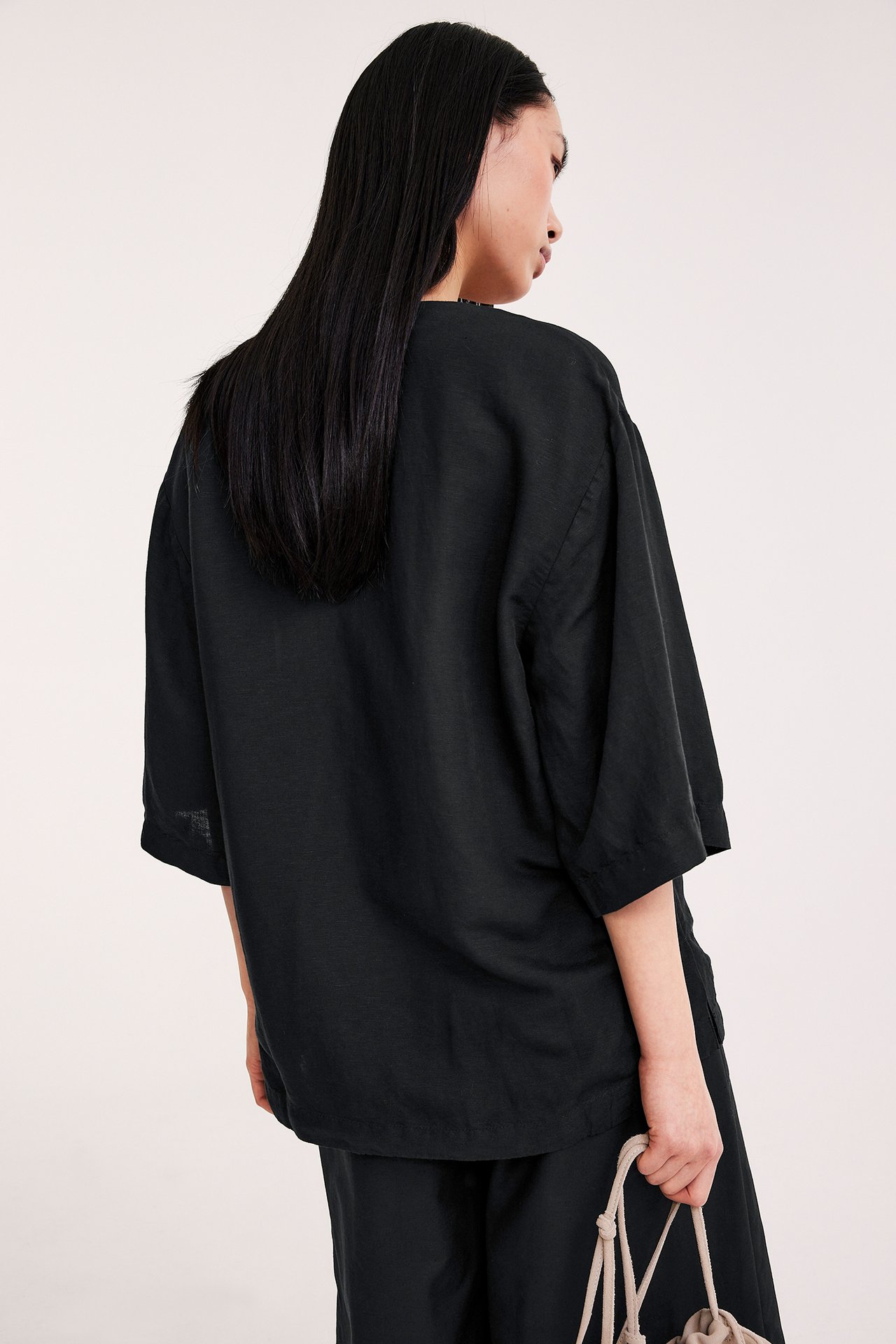 Ryggvy: kvinna i svart, lös kortärmad linneblandad skjorta, svarta byxor, håller beige väska.