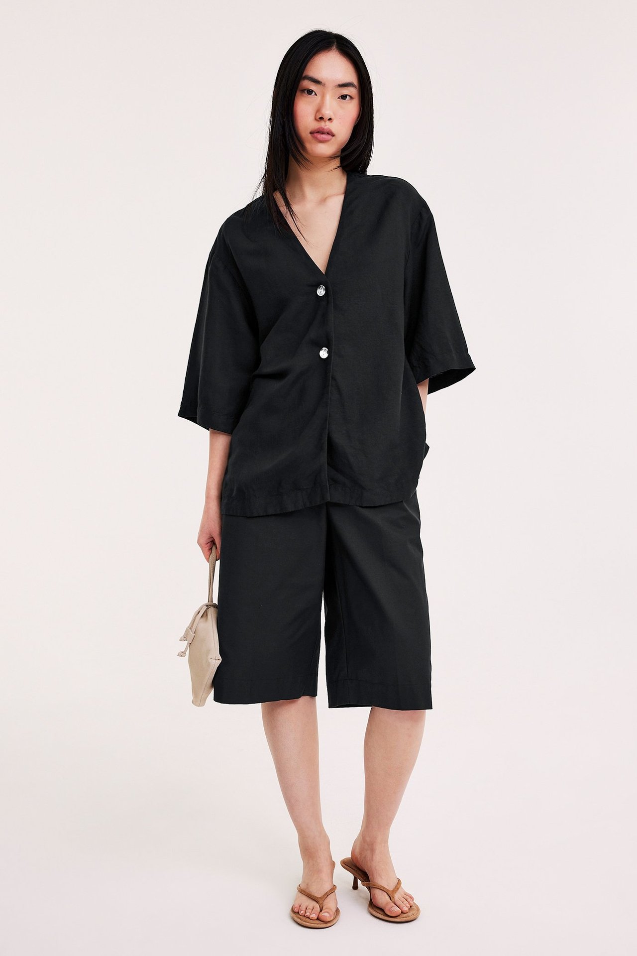 Kvinna i svart kortärmad skjorta i linnemix, svarta shorts, beige handväska och bruna klacktofflor.