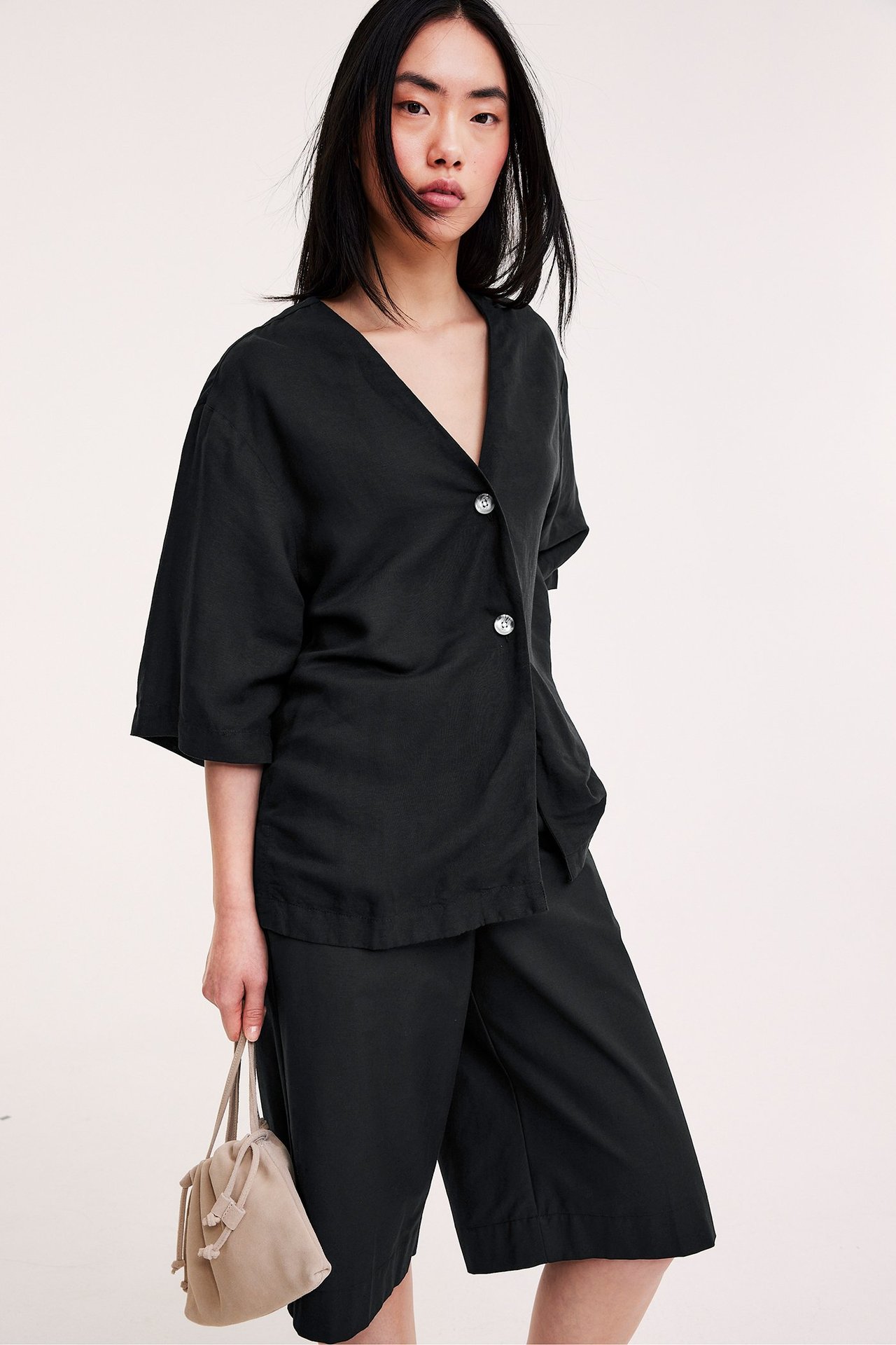 Edestä: Nainen mustassa löysässä V-pääntie-linsekoitepaidassa, kaksi valkoista nappia, mustat culotteshortsit ja beige laukku.