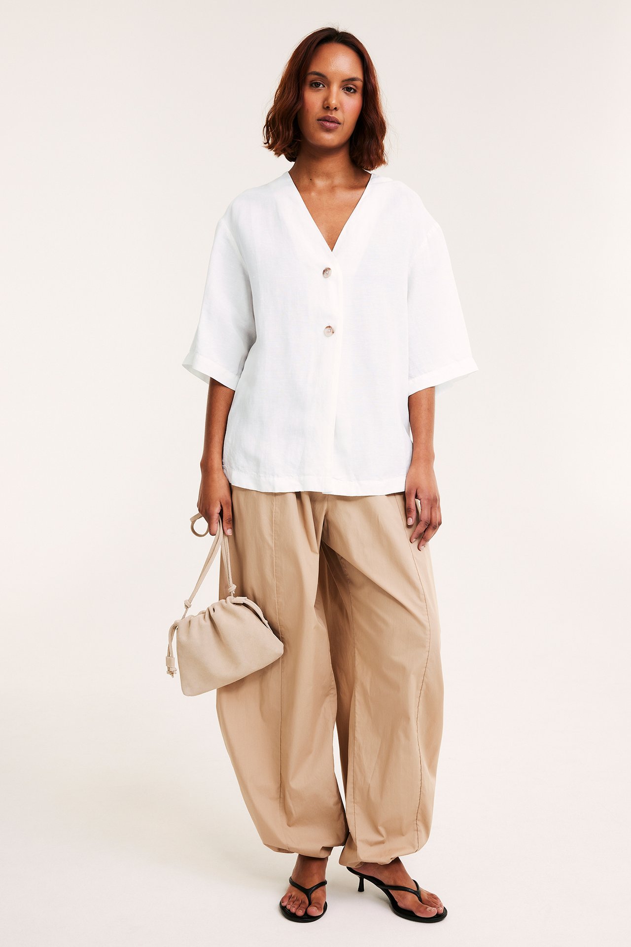 Kvinna bär vit kortärmad linneskjorta med knappar, beige vida byxor, svarta klacksandaler, håller beige dragskoväska.