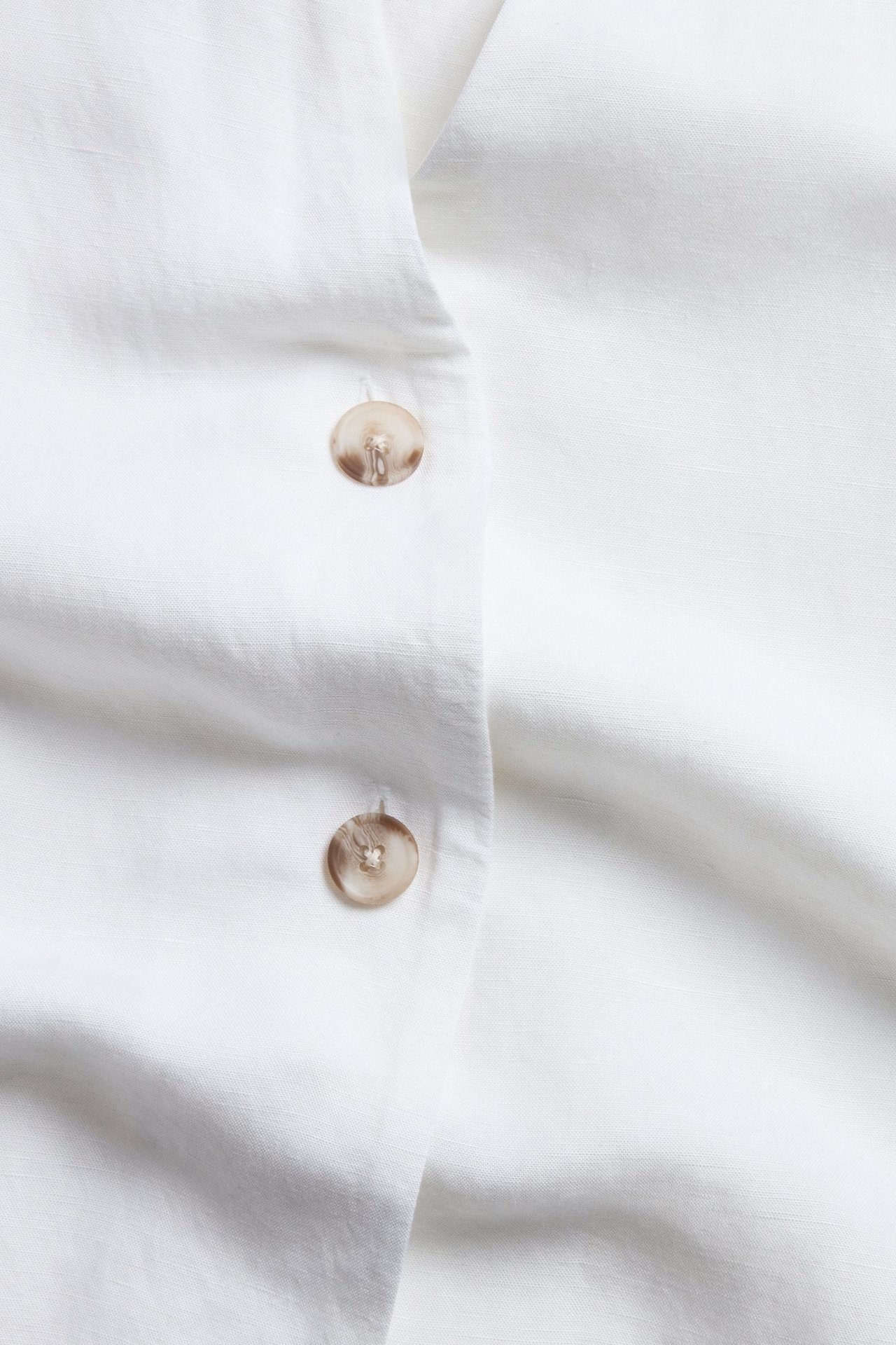 Zbliżenie białej koszuli z krótkim rękawem z mieszanki lnu, z dwoma jasnobrązowymi guzikami z przodu.