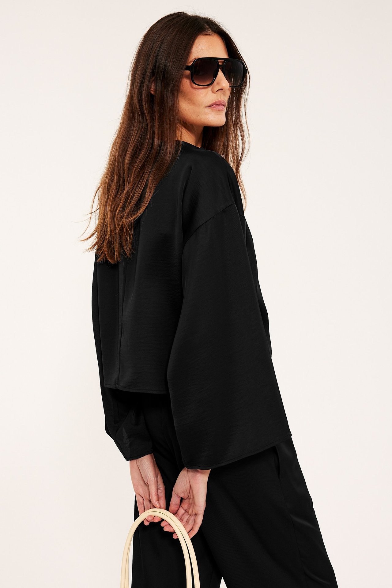 Sivukuva: nainen mustassa satiinipuserossa leveillä hihoilla, mustat housut, aurinkolasit, pitää vaaleaa laukkua.