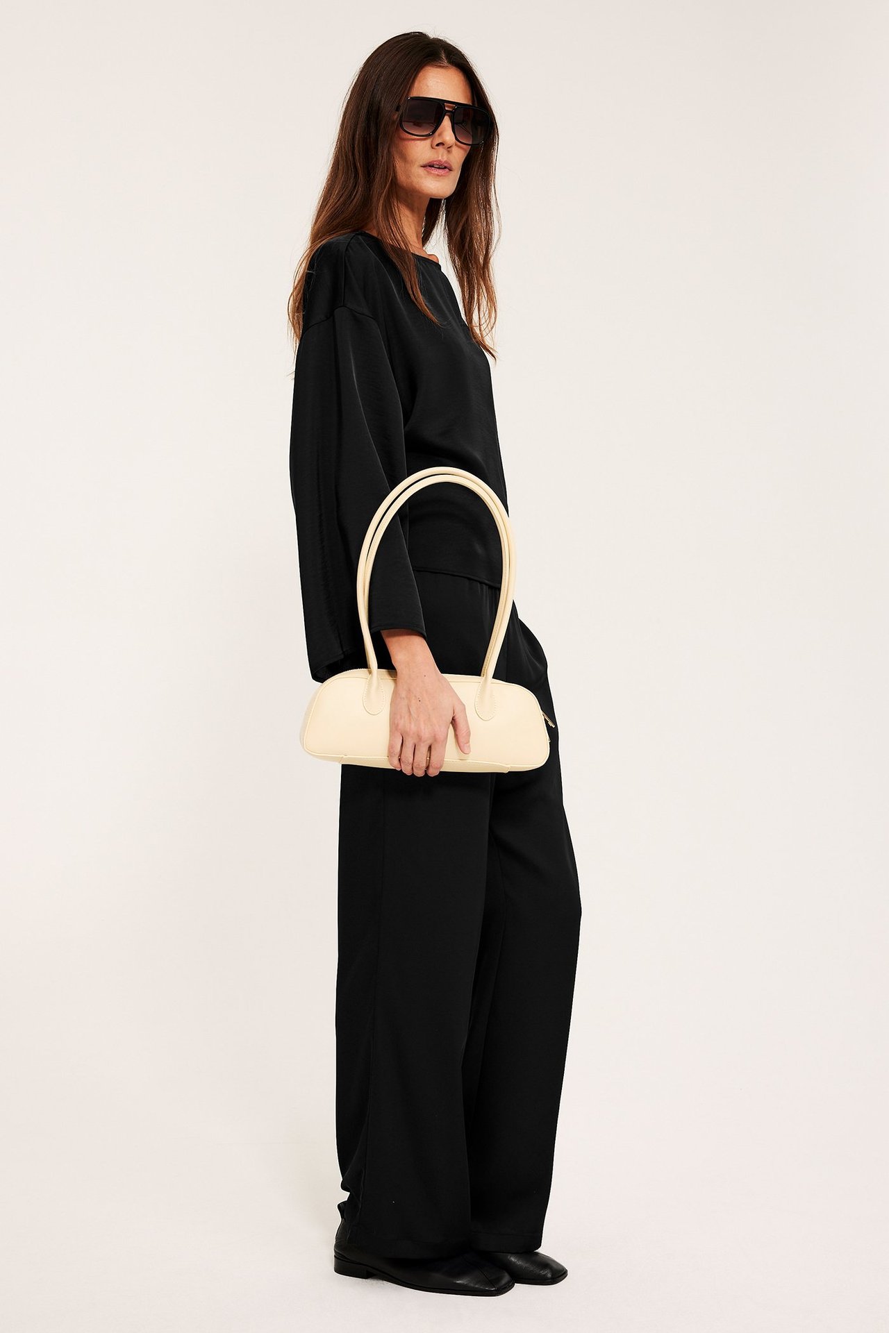 Naisen sivukuva mustassa satiinipuserossa, mustissa leveälahkeisissa housuissa, mustissa kengissä, aurinkolaseissa, pitäen kermanväristä käsilaukkua.