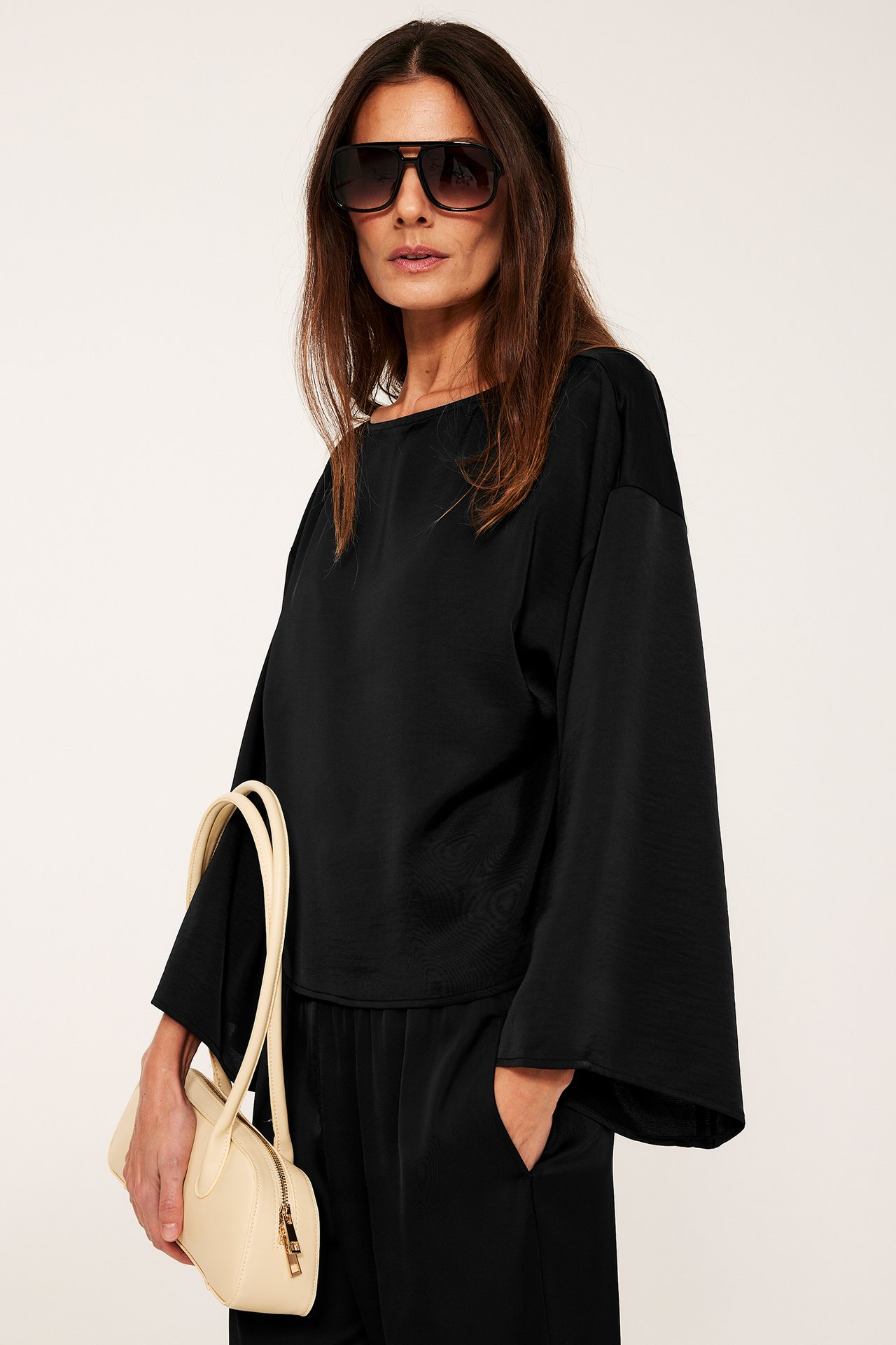 Nainen pukeutuu mustaan satiinipaitaan leveillä hihoilla, mustiin housuihin, aurinkolaseihin ja pitää beigeä käsilaukkua.
