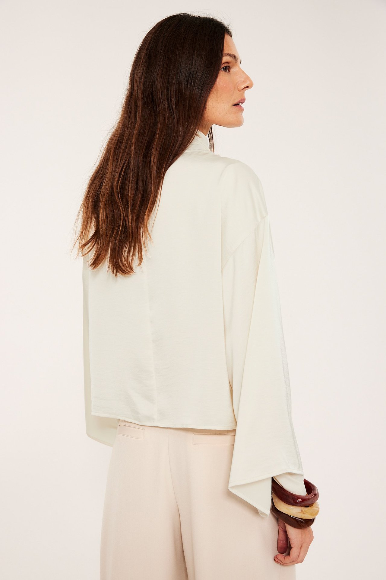 Naisen sivuselkäkuva offwhite satiinipuserossa, jossa leveät hihat, vaaleanbeiget housut ja rannekorut.