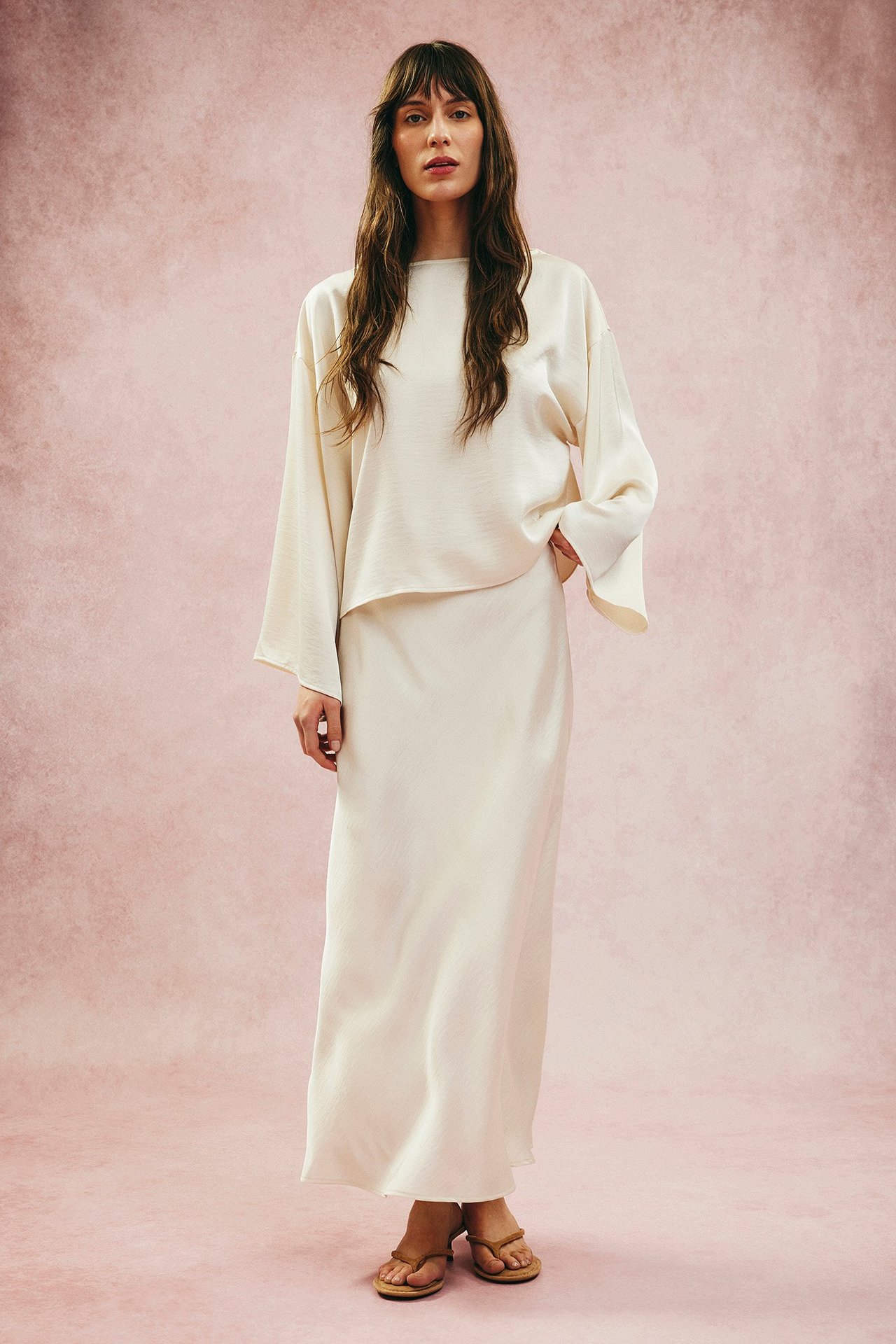 Z przodu: Kobieta w offwhite satynowej bluzce z szerokimi rękawami, długiej spódnicy i brązowych sandałach.