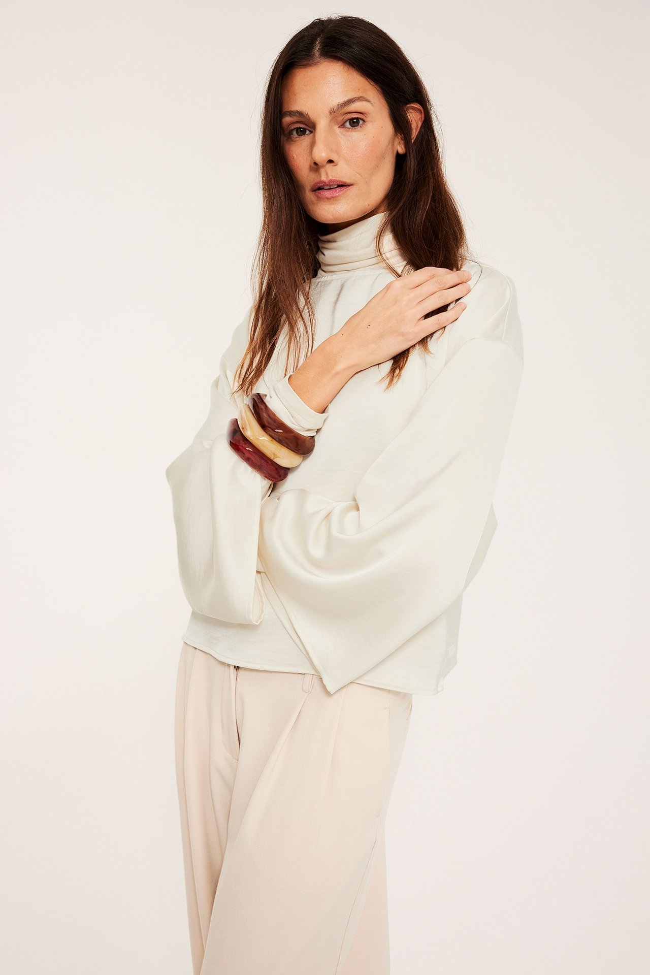 Edestä: Nainen luonnonvalkoisessa satiinipuserossa, jossa leveät hihat ja korkea kaulus, sekä vaaleat housut.