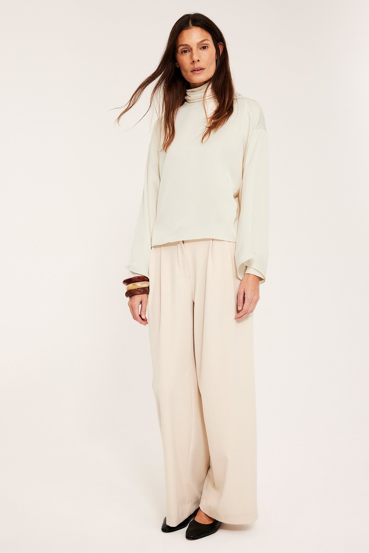 Edestä: Nainen offwhite satiinipuserossa leveillä hihoilla, vaaleat leveät housut, ruskeat rannekorut ja mustat kengät.