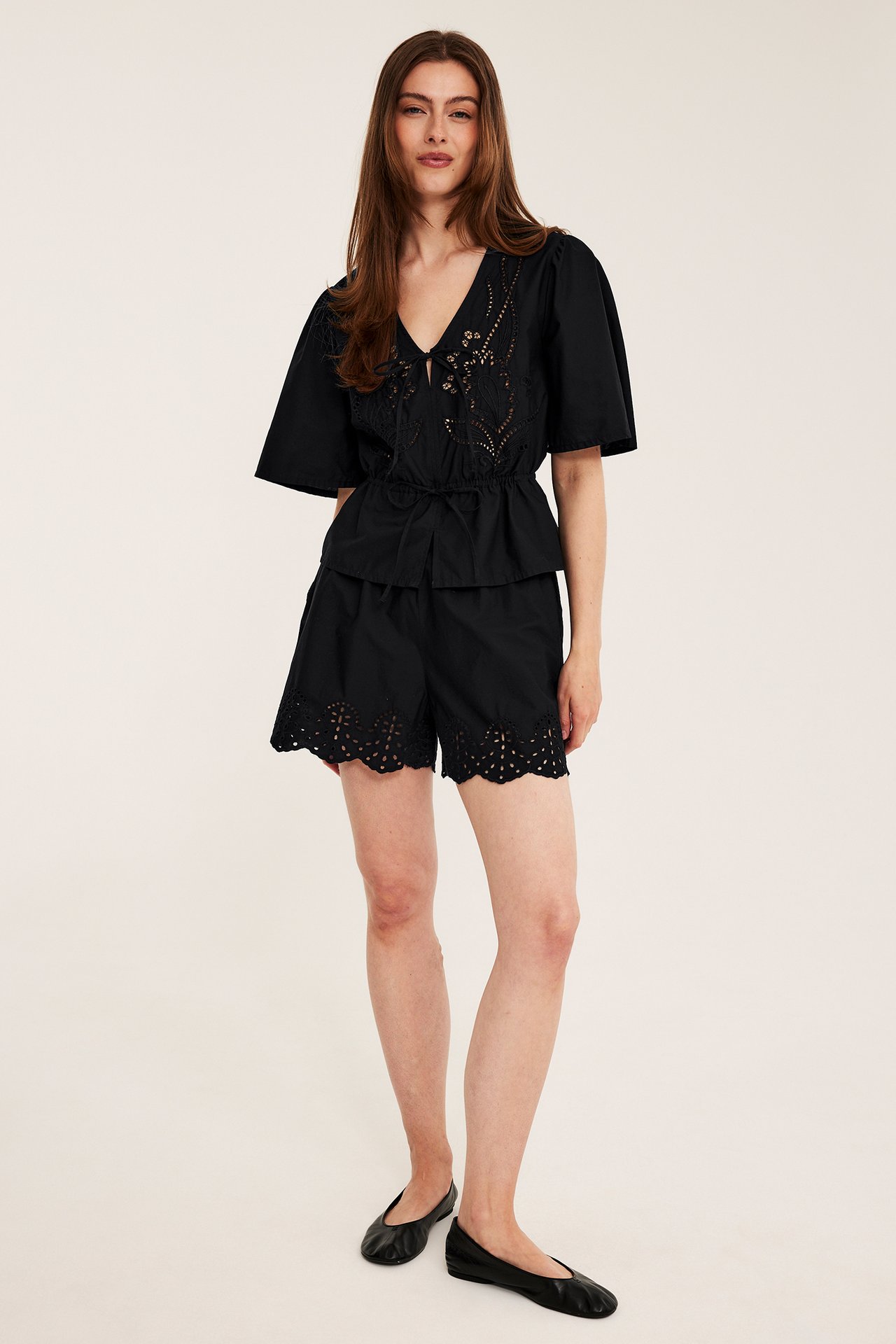 Frontvy av kvinna i svart broderad bomullspoplinblus med midjeband, matchande shorts och svarta ballerinaskor.