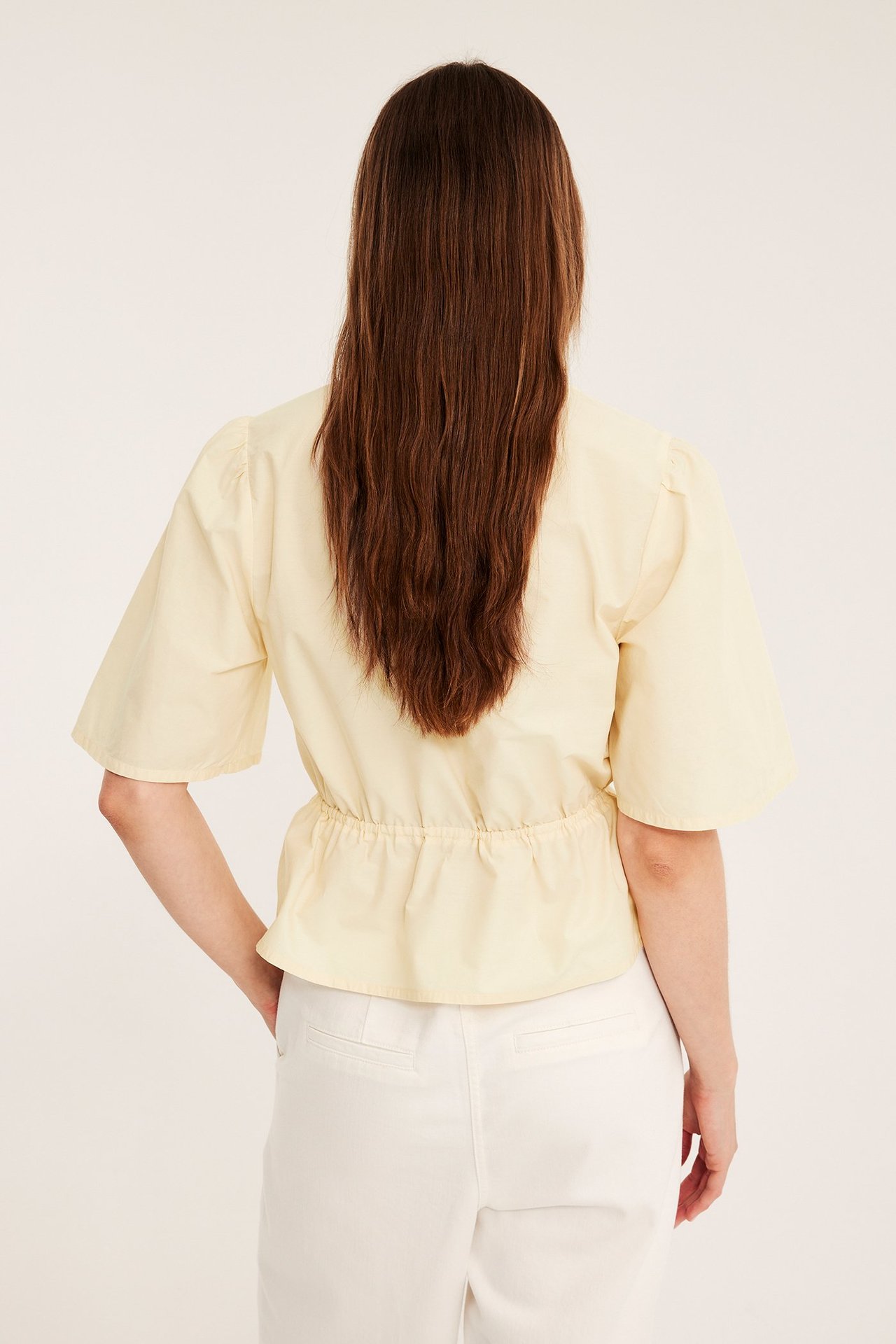 Bakfra: kvinne i lys gul poplinbluse med puffermer, peplum-midje og hvite bukser.