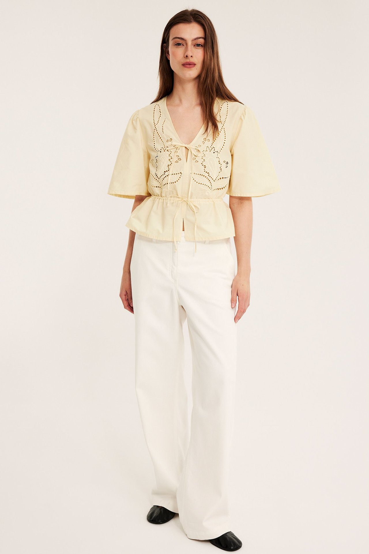 Edestä: Nainen yllään vaaleankeltainen kirjailtu puuvillapopliinipusero vyötärönauhoilla, valkoiset leveälahkeiset housut, mustat kengät.