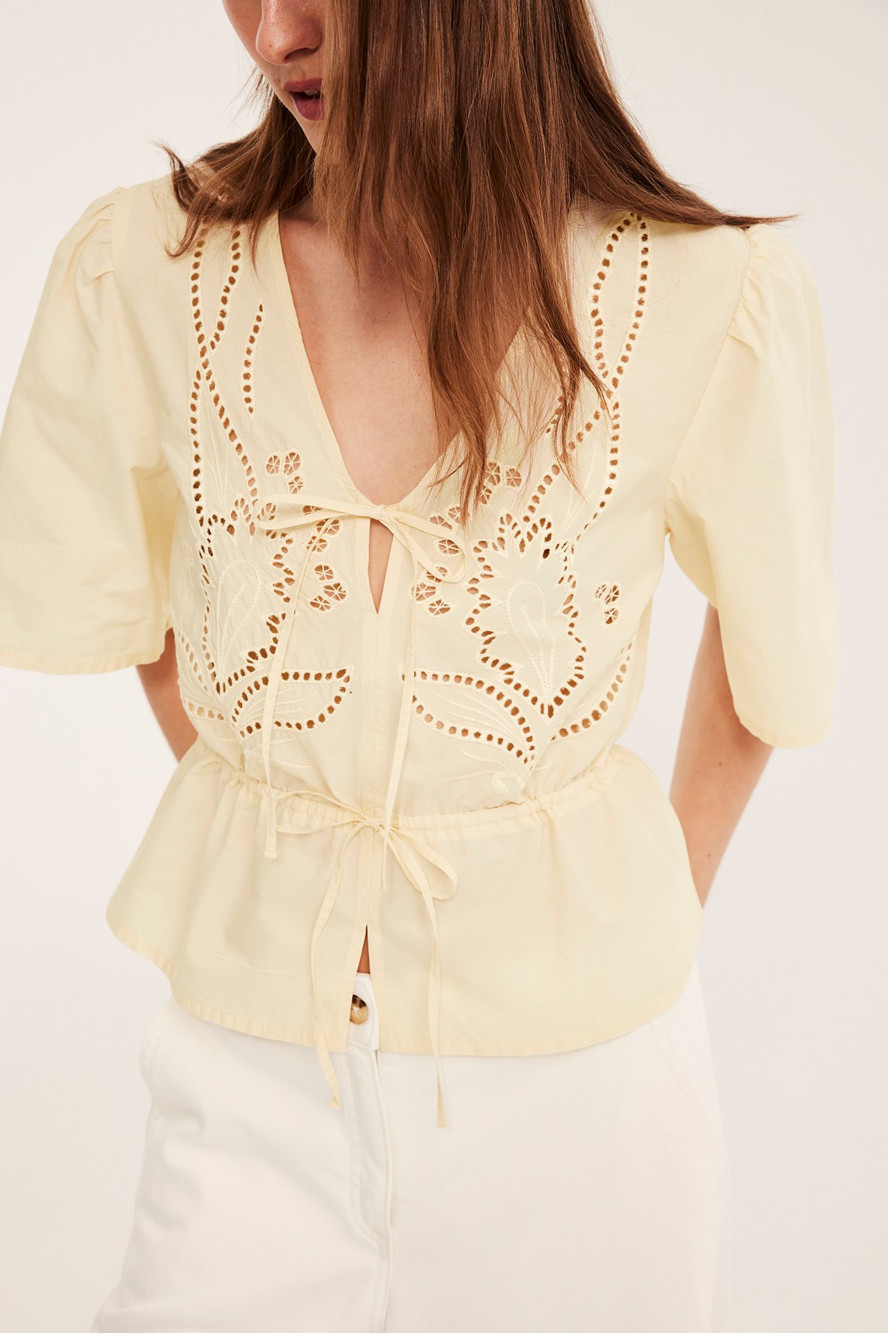 Naisen etukuva, yllään vaaleankeltainen puuvillapopliinipusero valkoisella kirjailulla, vyötärönauhalla ja valkoiset housut.