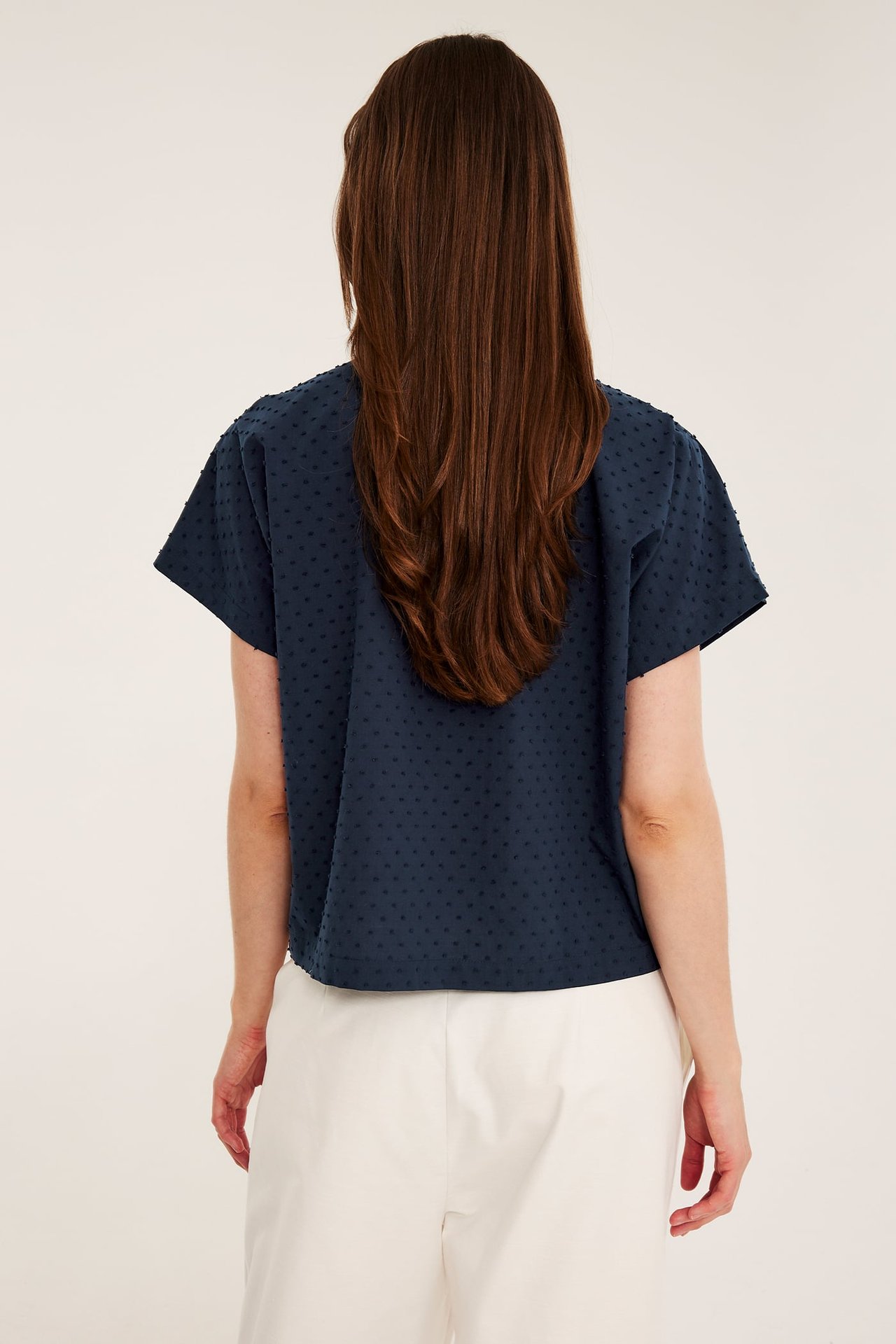 Widok z tyłu kobiety w granatowej, kropkowanej bawełnianej koszuli z krótkim rękawem i jasnych luźnych spodniach.