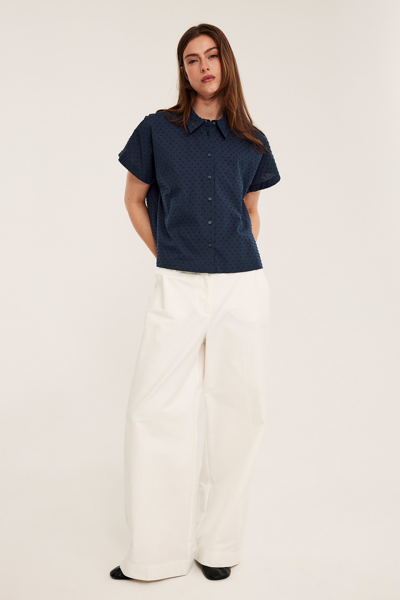 Framifrån, kvinna i mörkblå prickig kortärmad bomullsskjorta, vita vida byxor och svarta skor.