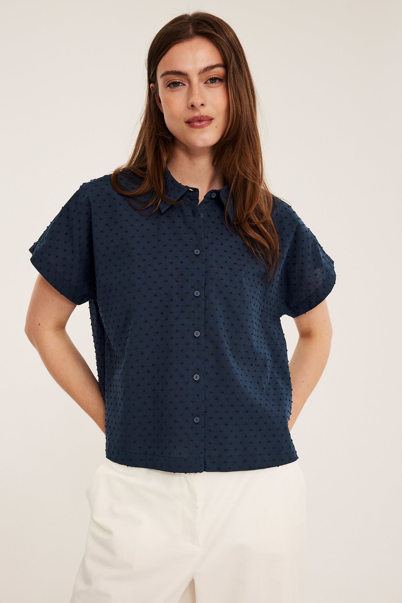 Framifrån: Kvinna i mörkblå prickig kortärmad bomullsskjorta med krage och vita byxor.