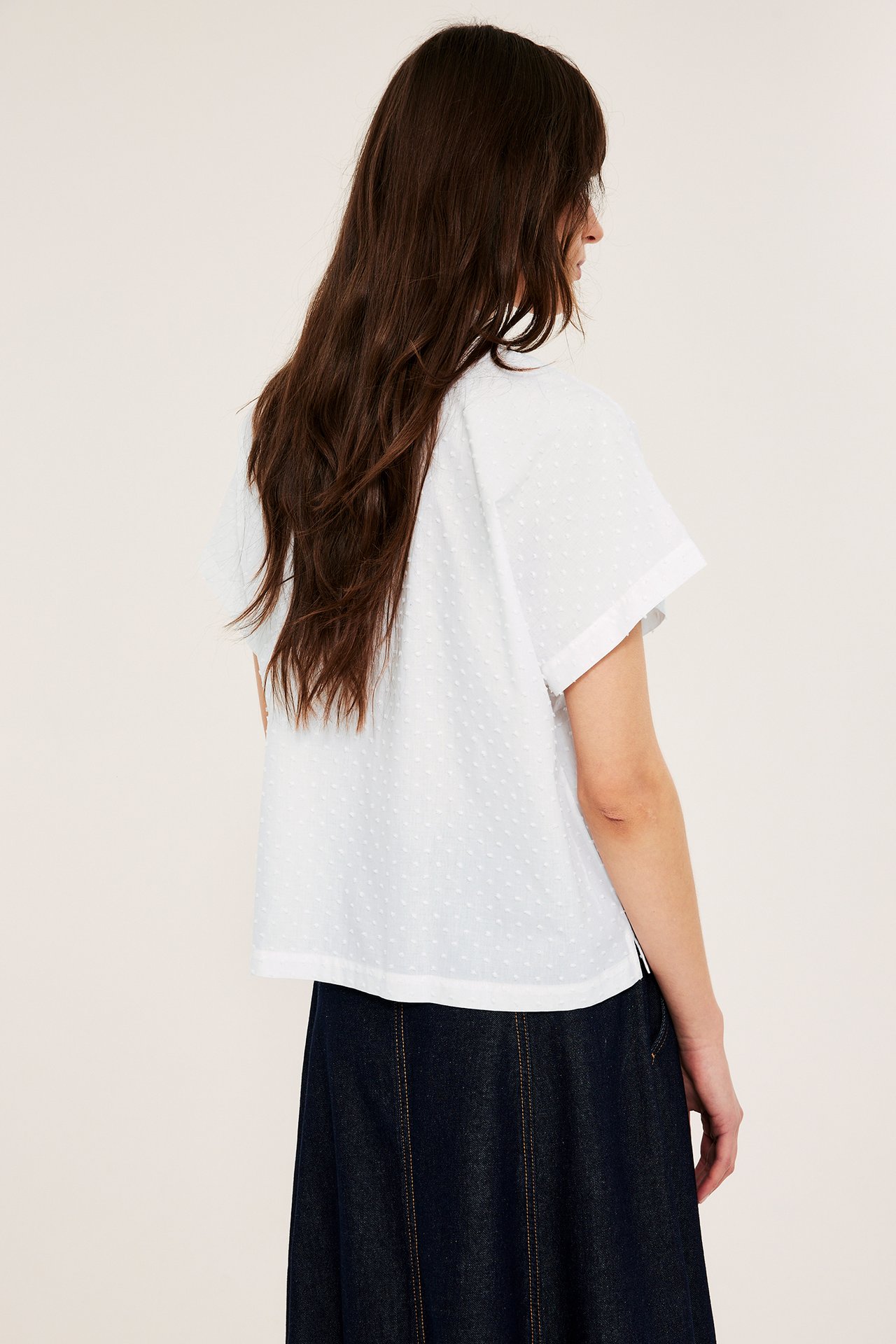 Bakvy av en kvinna i en vit kortärmad prickig bomullsskjorta och en mörkblå denim A-linjekjol.