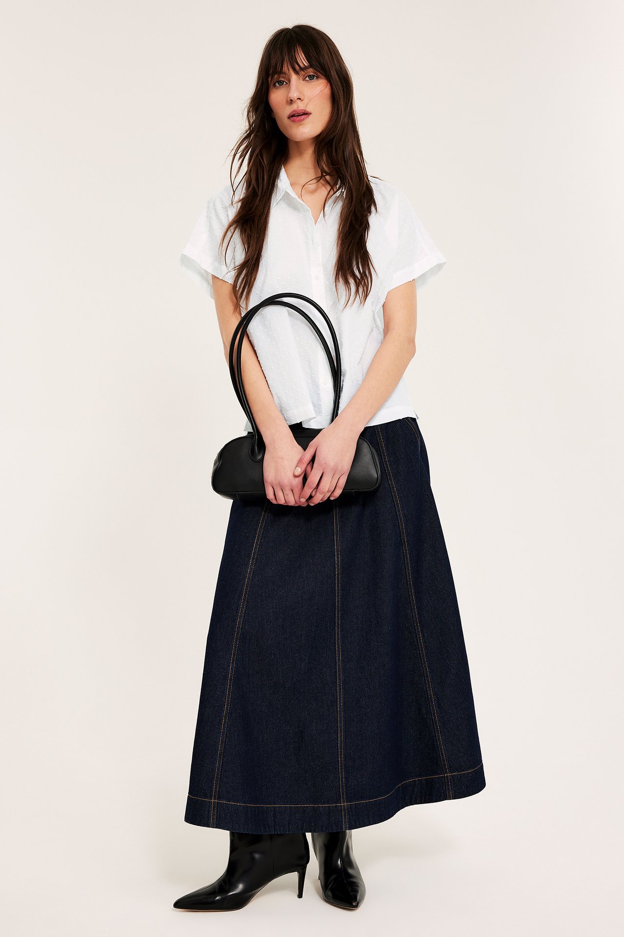 Framifrån på en kvinna i en vit kortärmad prickig bomullsskjorta, mörkblå jeanskjol, svarta stövlar och med en svart handväska.