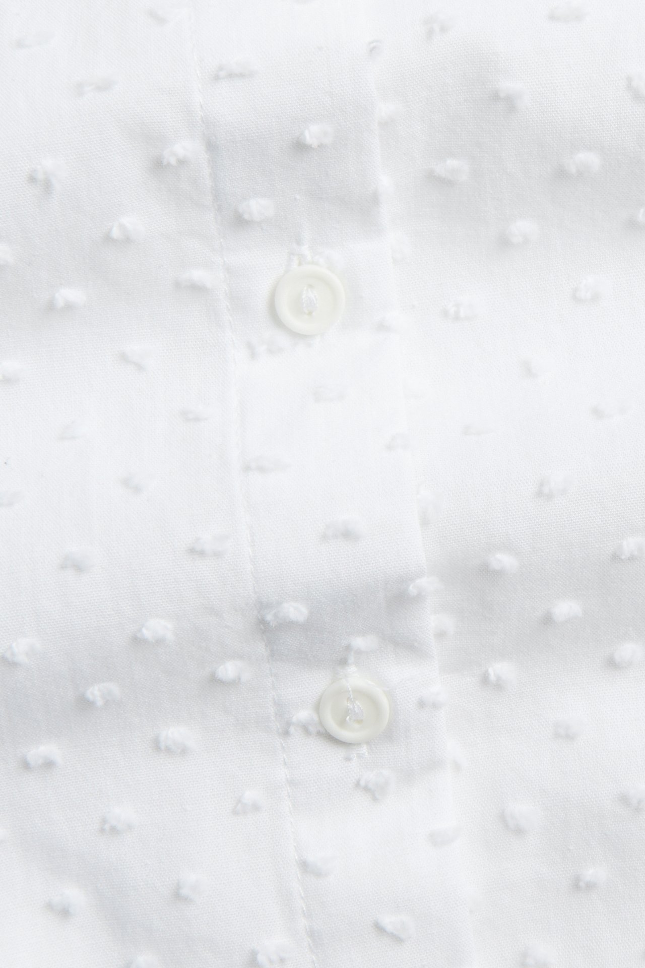 Närbild av en vit bomullsskjorta med upphöjda prickar och två vita knappar på en knappslå.