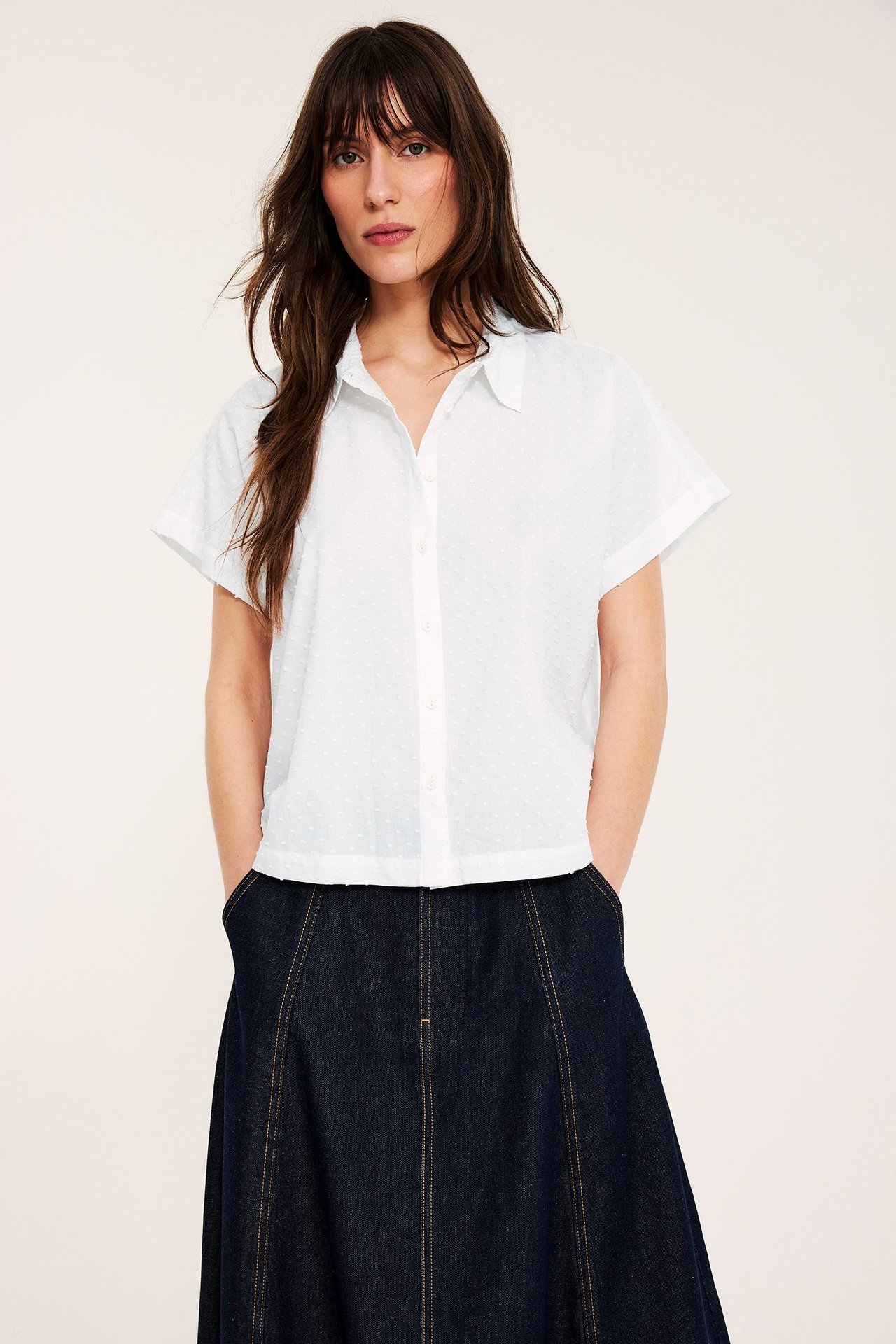 Frontvy av en kvinna i vit kortärmad prickig bomullsskjorta med krage och mörkblå jeanskjol.