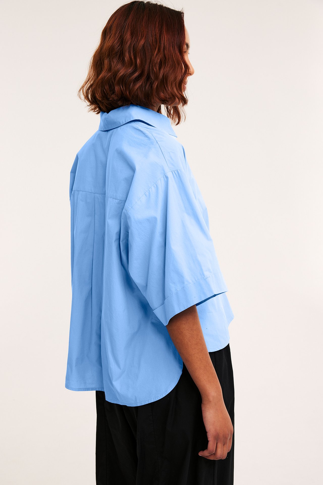 Sidebilde av en kvinne i en løs lys blå kortermet skjorte i bomullspoplin og svarte bukser.