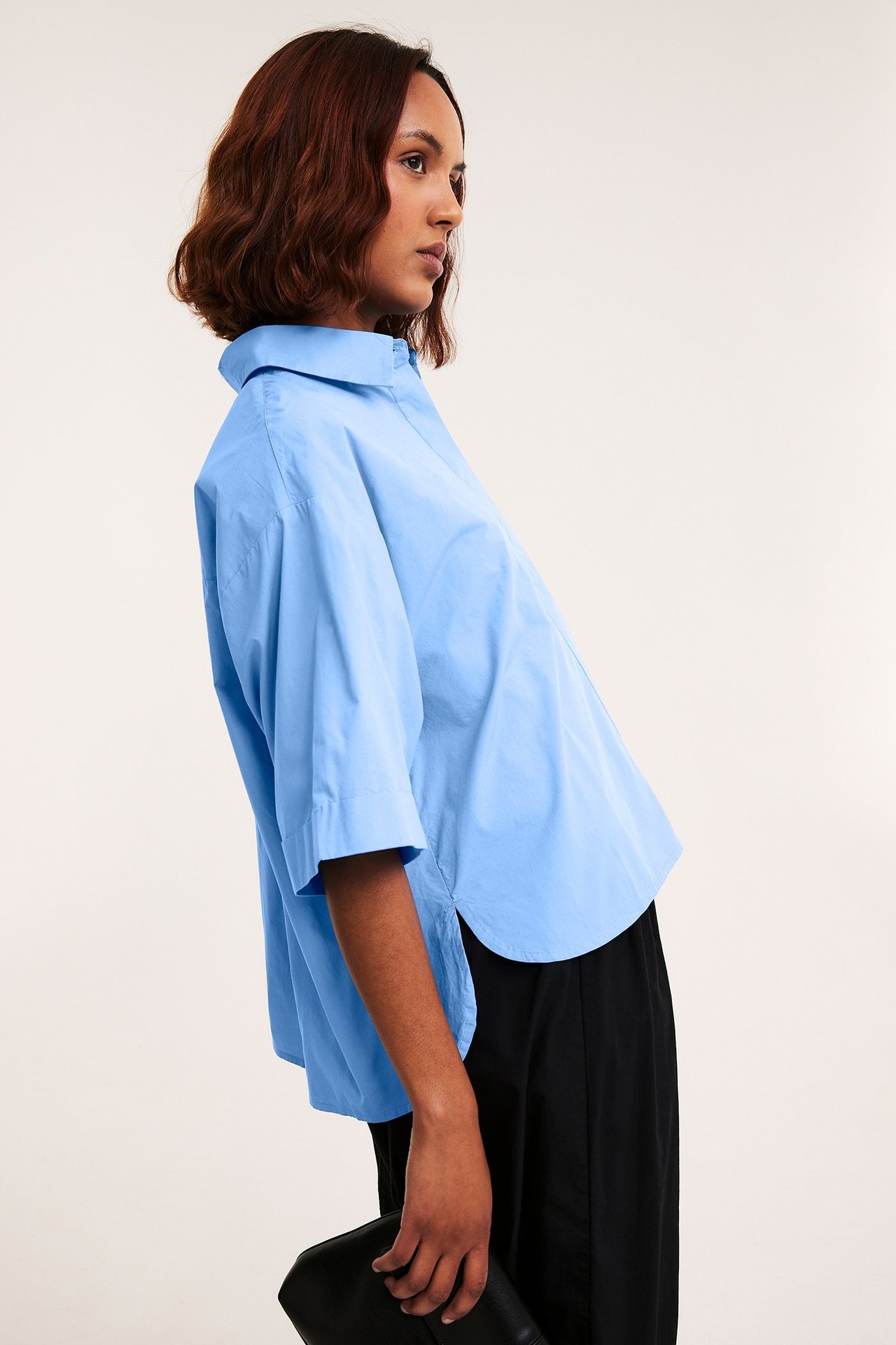 Sidebilde: kvinne i lyseblå kortermet poplinskjorte, svarte bukser, holder svart clutchveske.