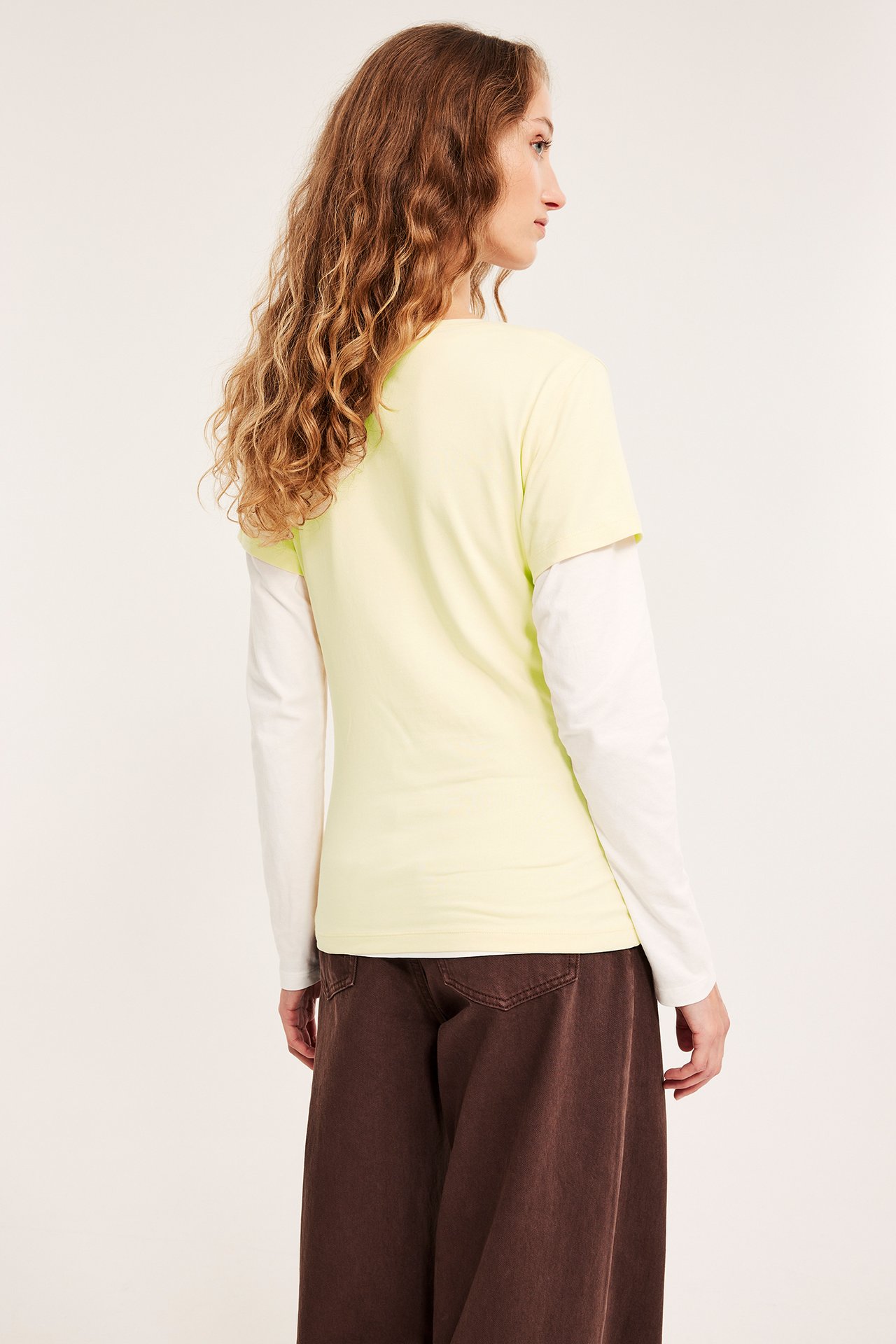 Kobieta, tyłem, w jasnożółtym topie, białej bluzce z długim rękawem i brązowych spodniach.