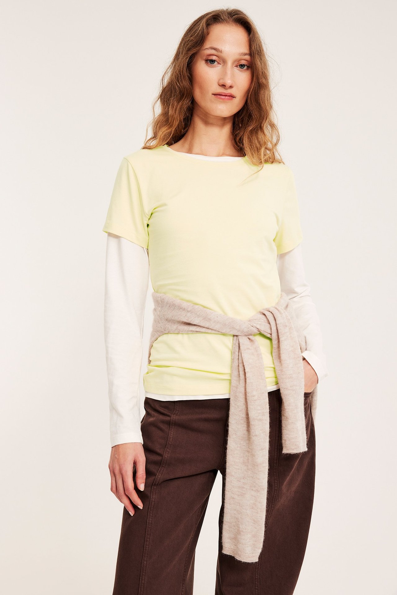 Widok z przodu: Kobieta w jasnożółtym topie z krótkim rękawem na białej koszuli z długim rękawem, brązowe spodnie, beżowy sweter w pasie.