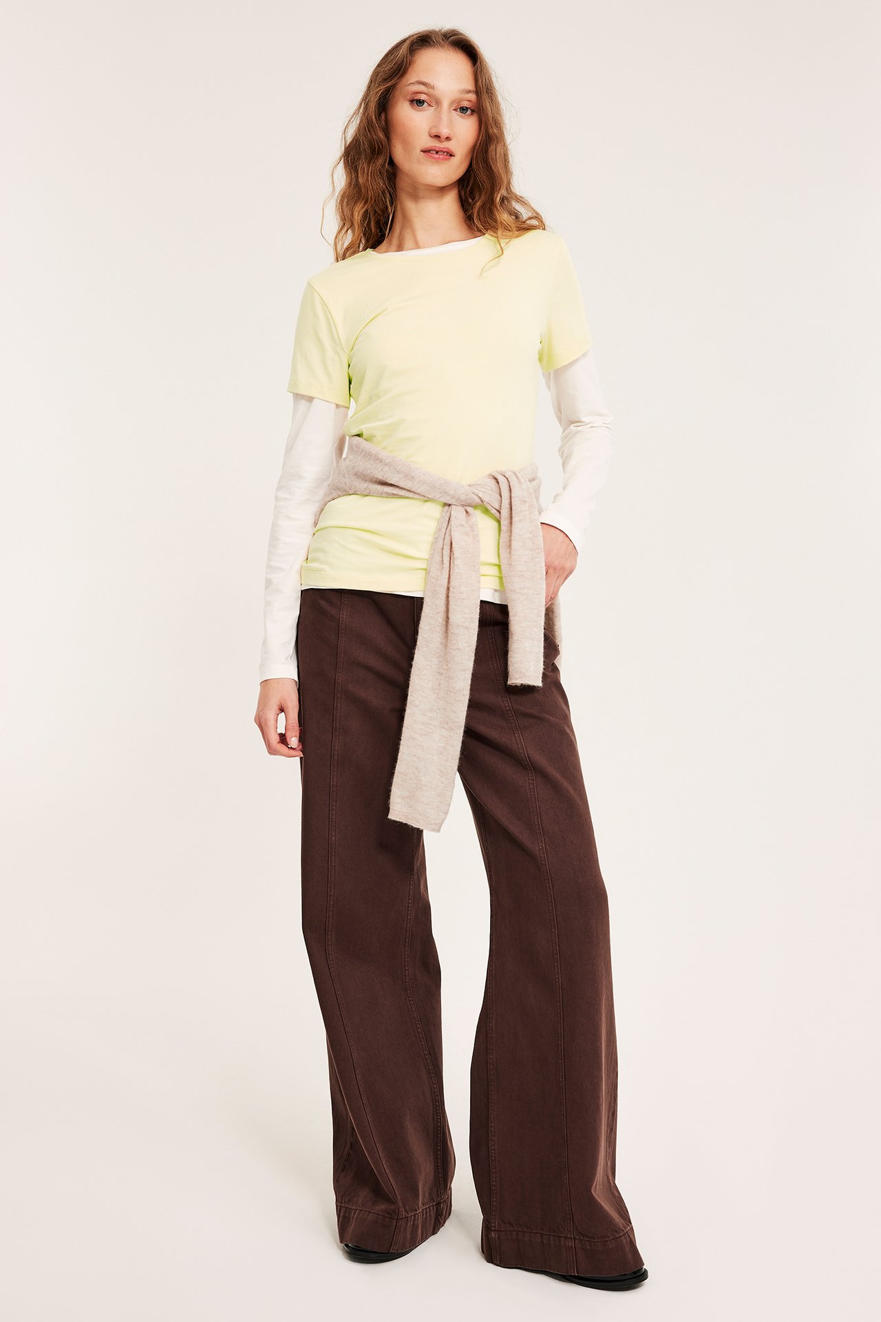 Widok z przodu: Kobieta w jasnożółtym topie z krótkim rękawem na białym długim rękawie, brązowych szerokich spodniach, beżowy sweter w pasie.