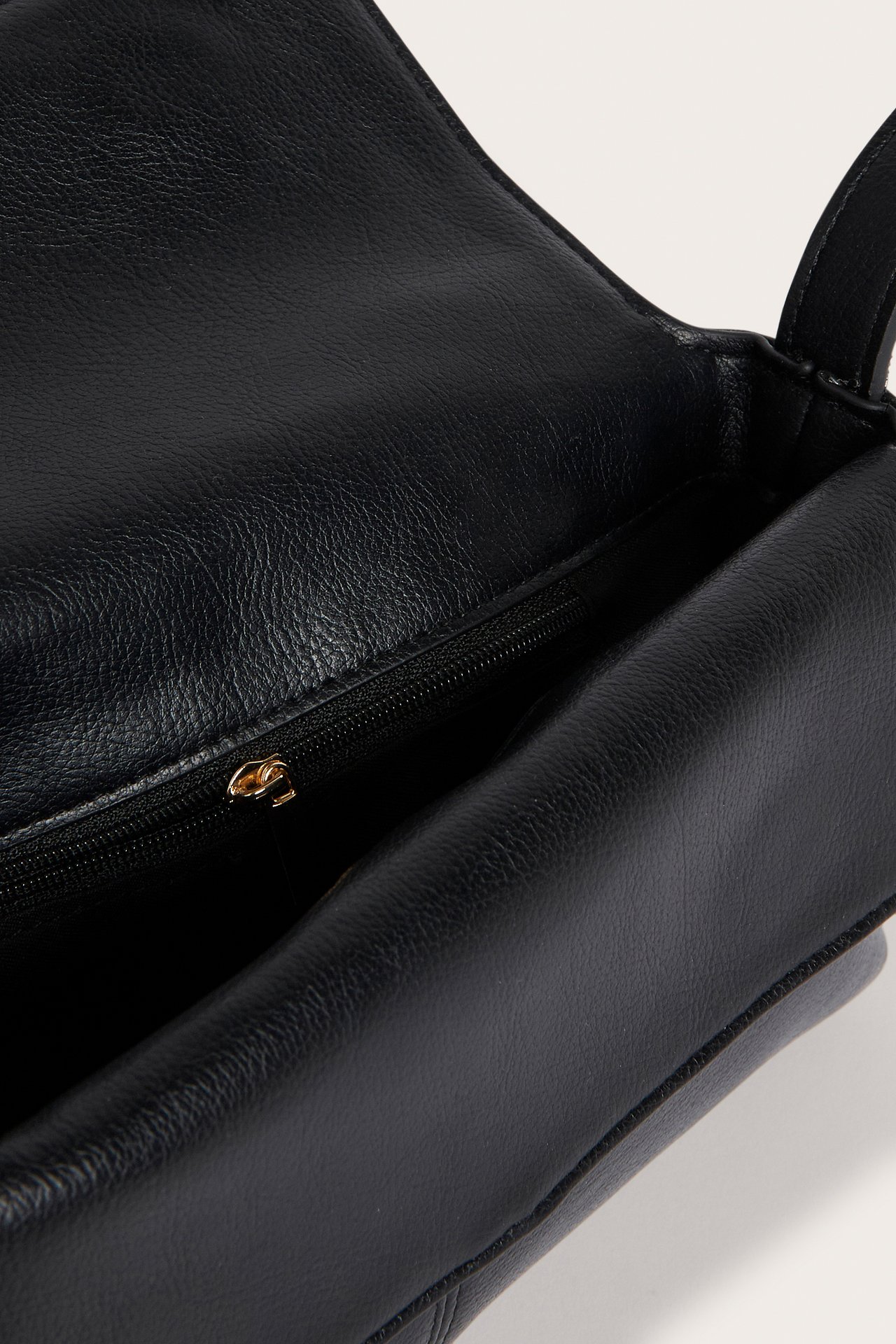 Nærbilde av en åpen svart skulderveske i imitert skinn, som viser den indre glidelåslommen med gullfarget glidelås.