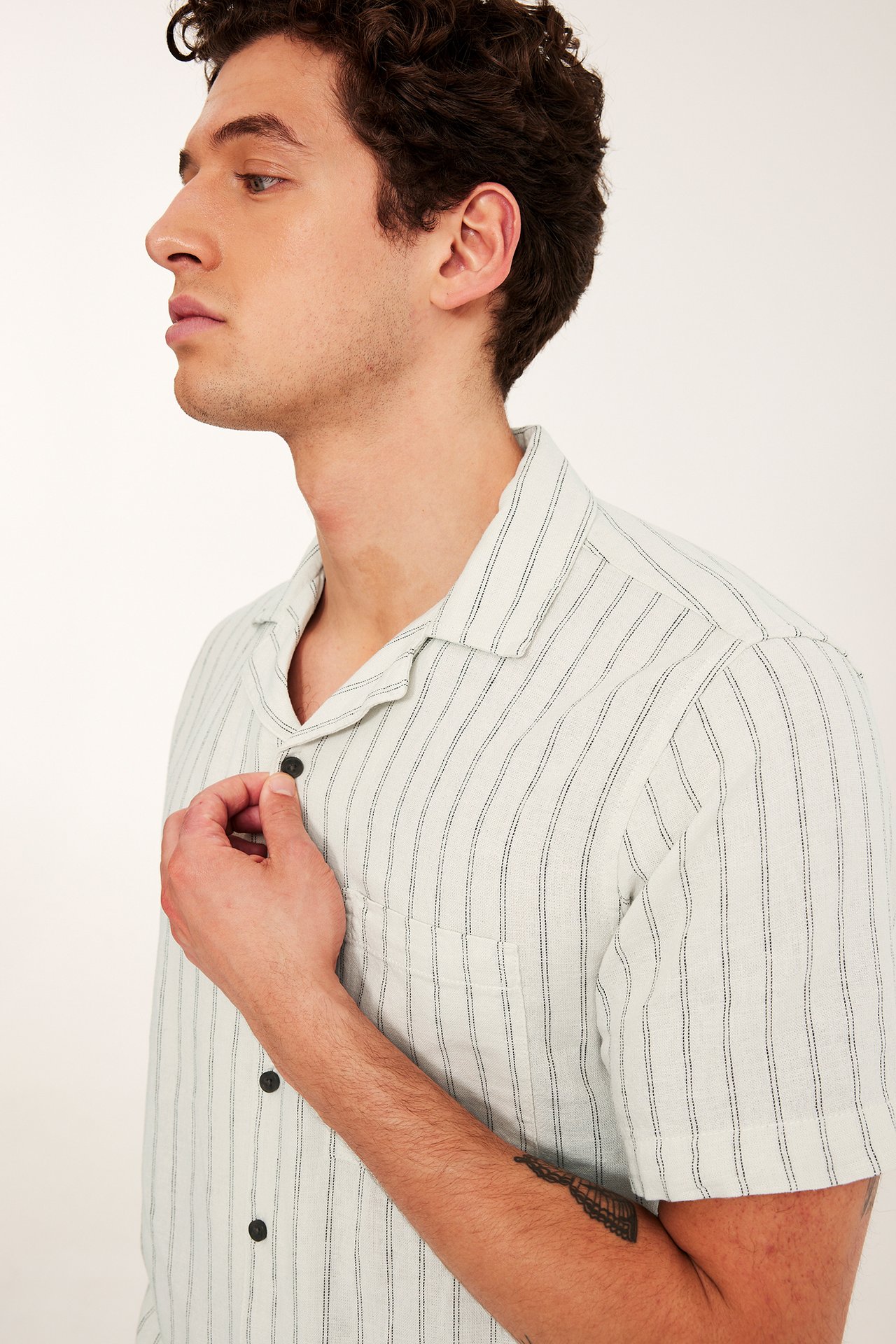 Zbliżenie, widok z boku: Mężczyzna w jasnobeżowej lniano-wiskozowej koszuli resortowej w paski, ciemne guziki.