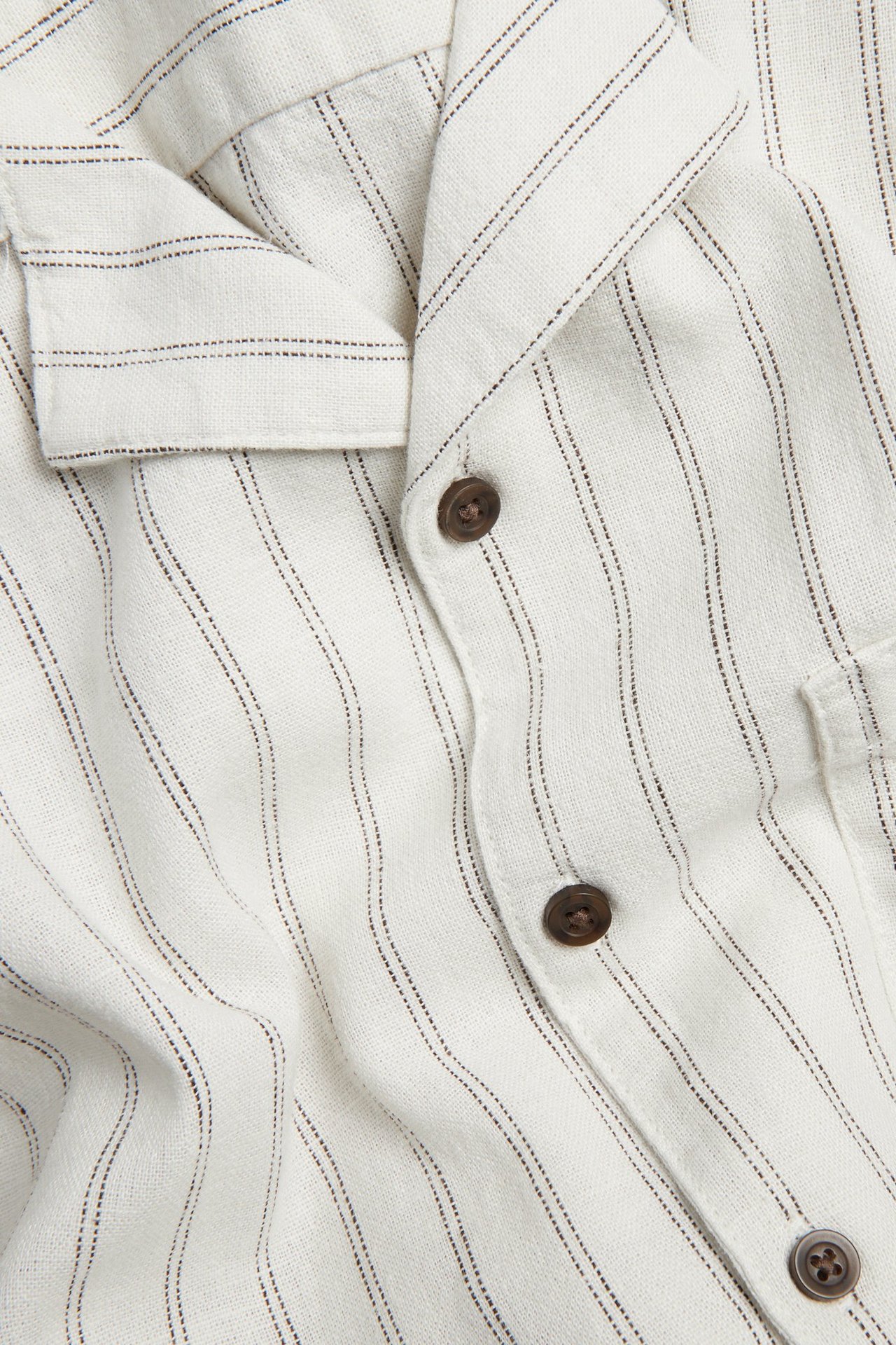Nærbilde av lys beige stripete herreskjorte i lin-viskosemix med resortkrage og brune knapper.