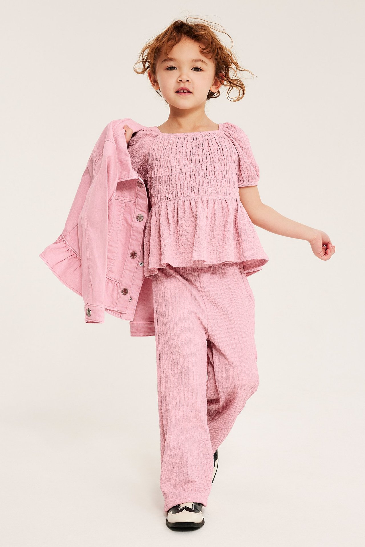 Front view av ett barn i ljusrosa smockad peplumtopp, matchande texturerade byxor, håller rosa jeansjacka.
