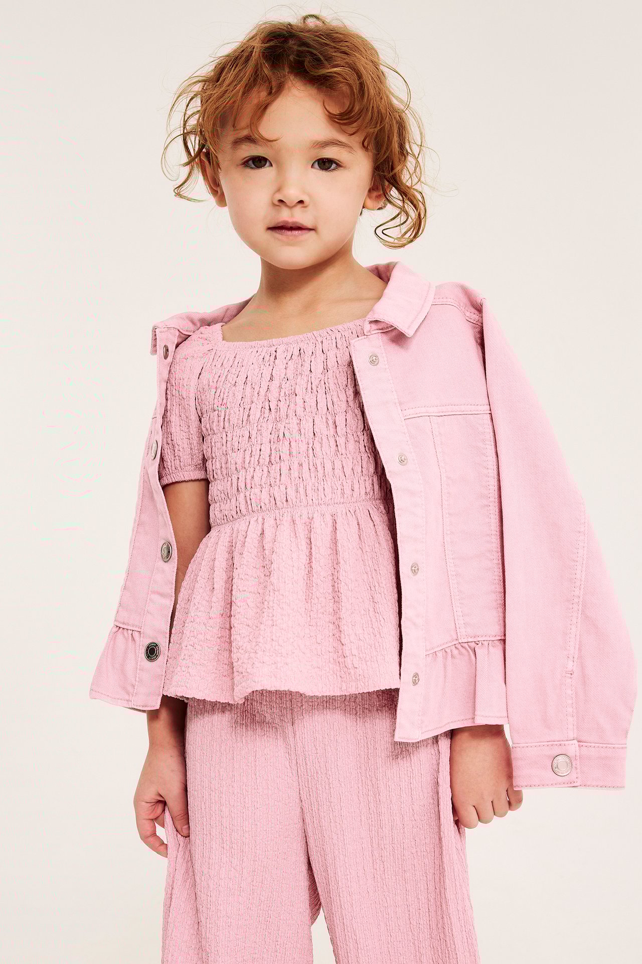 Widok z przodu: Dziecko w jasnoróżowym marszczonym topie, różowej kurtce jeansowej i szerokich spodniach.