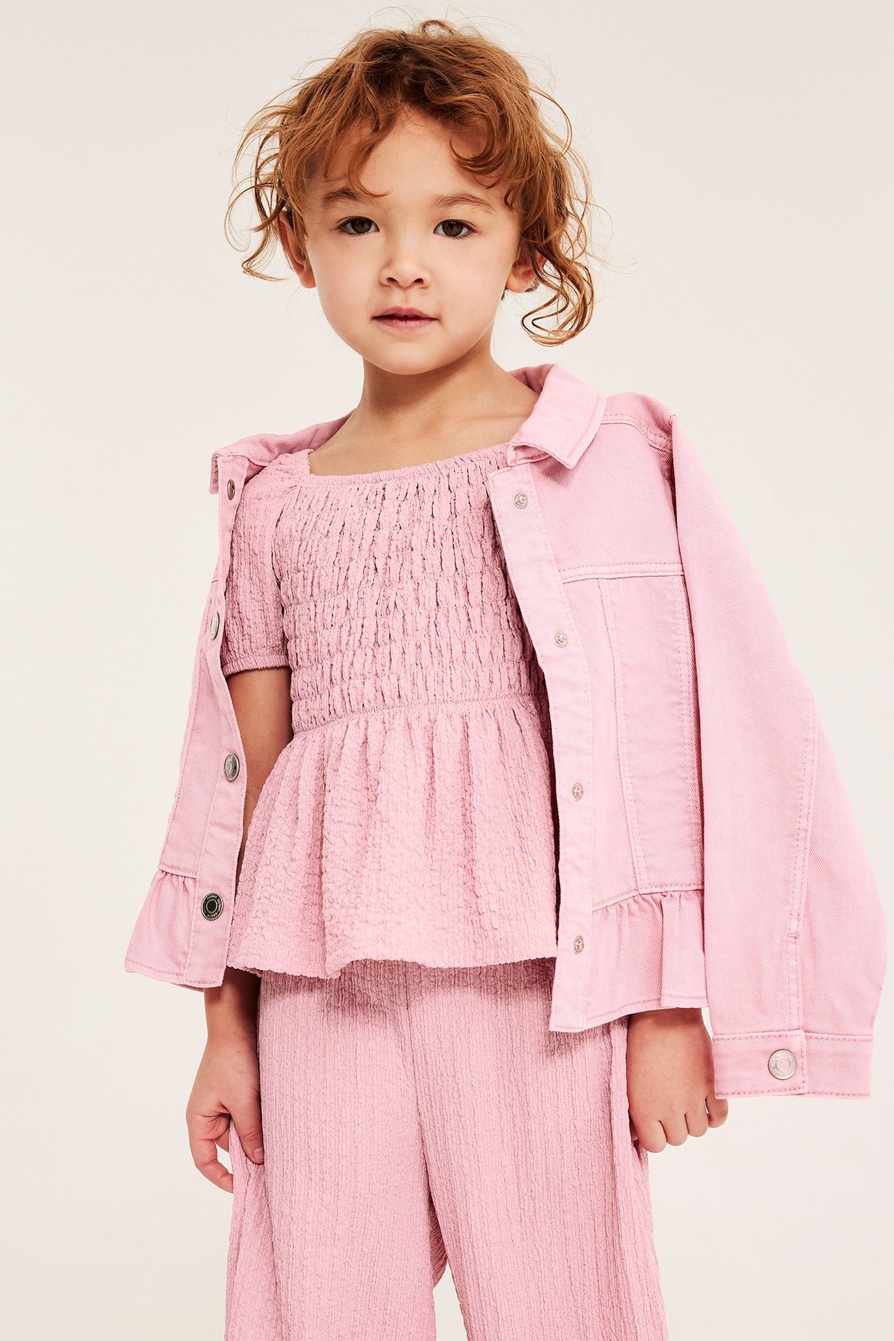 Frontvy: Ett barn bär en ljusrosa krinklad smocktopp, rosa jeansjacka och vida byxor.